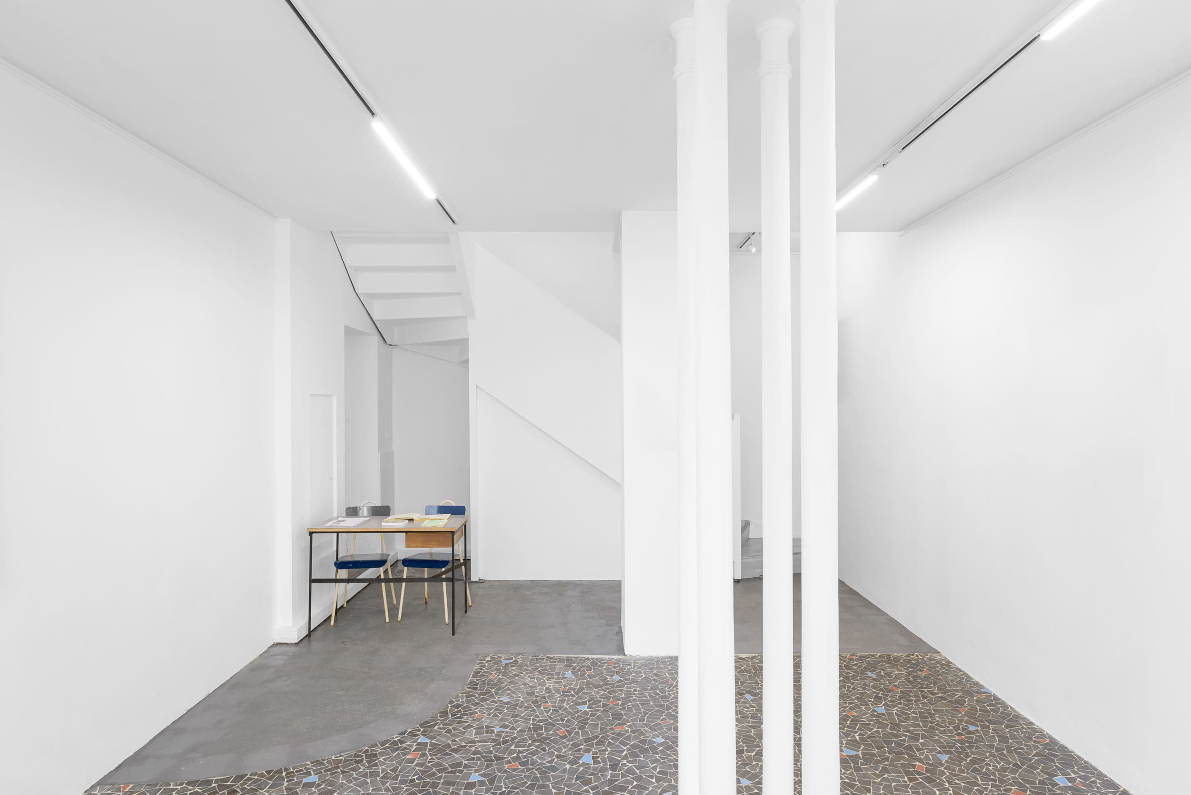 The image shows a minimalist interior space featuring white walls, a staircase, a table with chairs, and a patterned floor.
