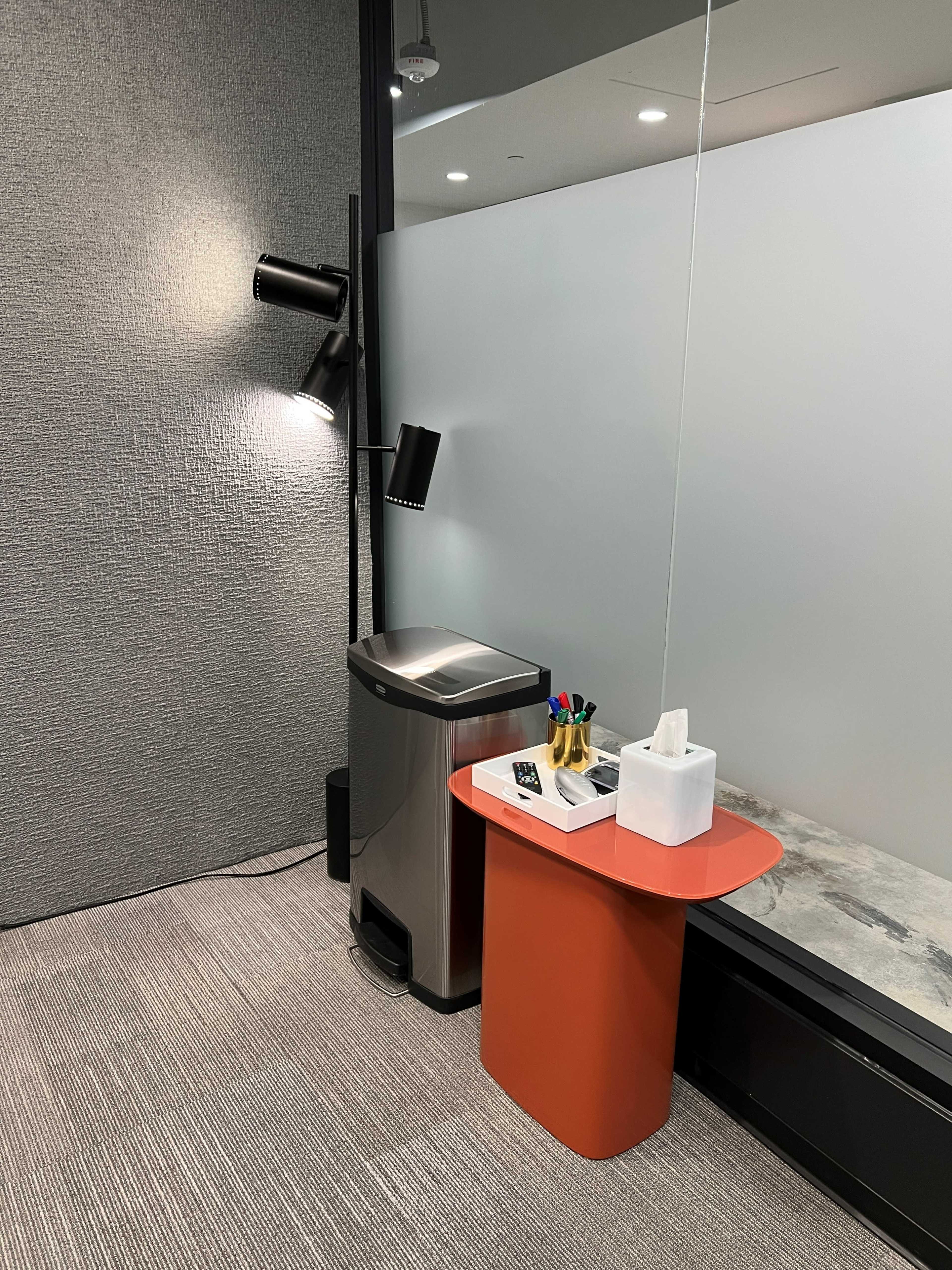 A modern office corner with a gray wall, a trash can, a small round orange table, and a set of office supplies on the table.