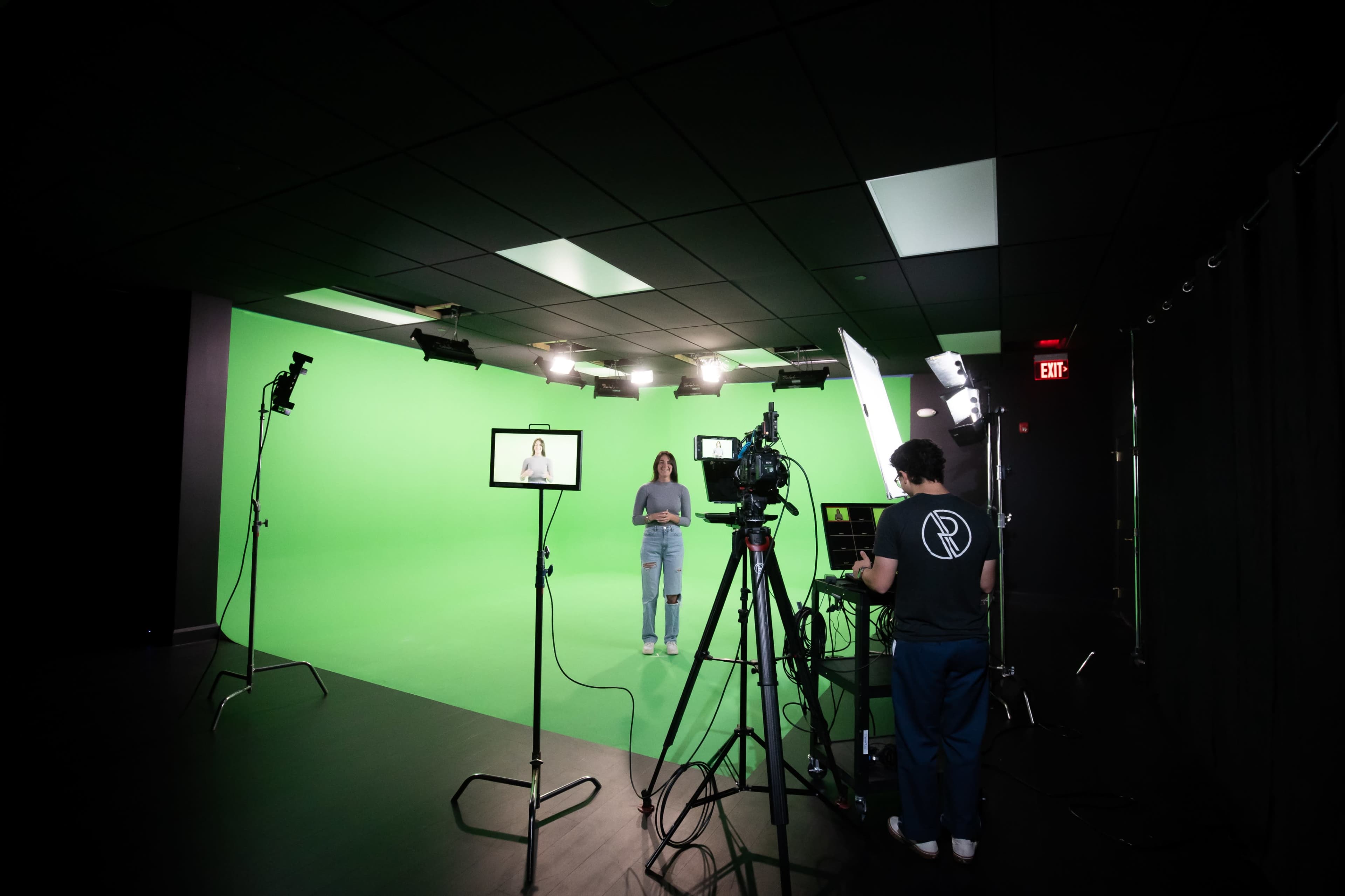 A person stands in front of a green screen while another individual operates equipment and cameras in a studio setting.