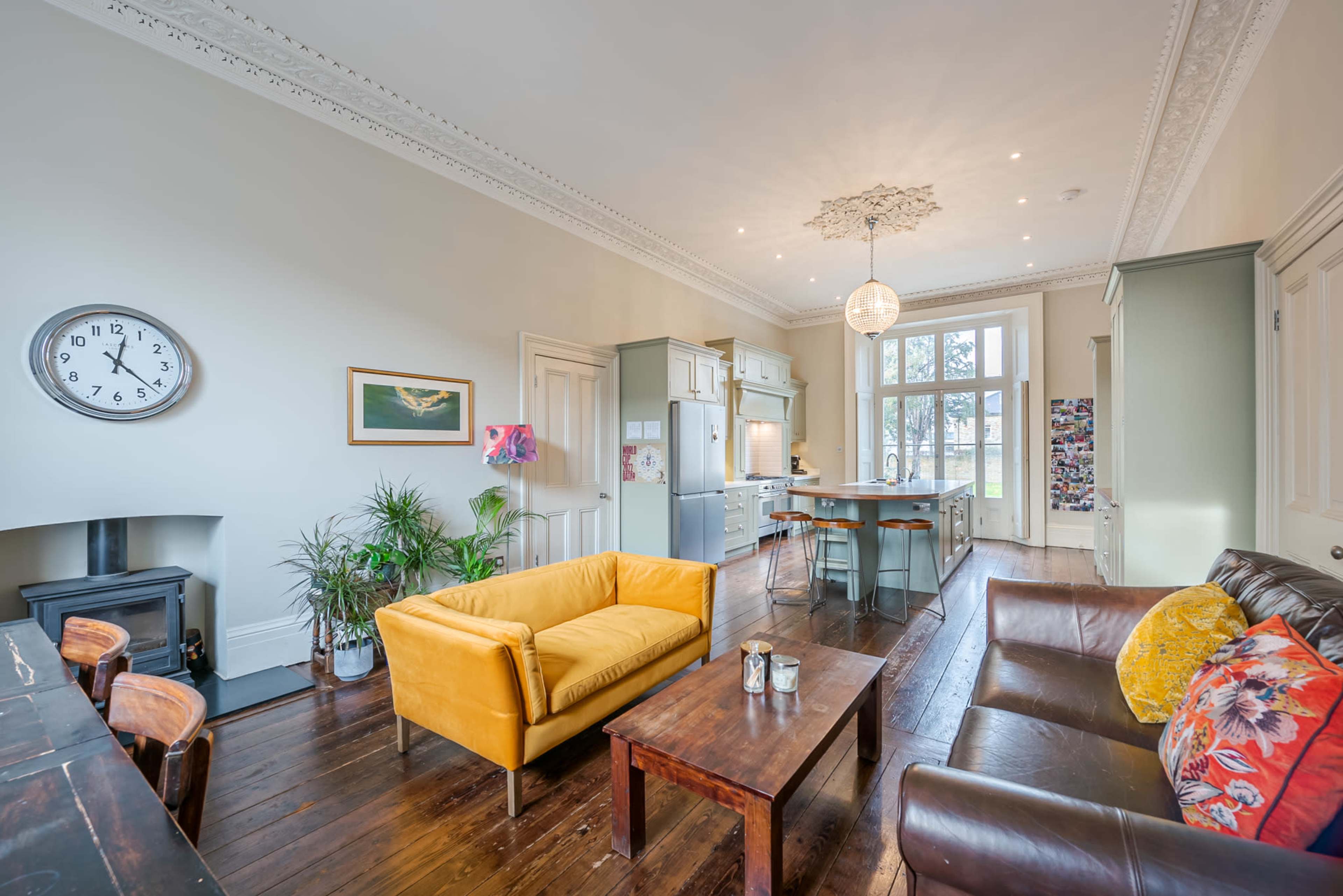 A stylish living room with a yellow sofa, a brown leather couch, and a wooden coffee table, leading to a kitchen area with large windows and modern appliances.