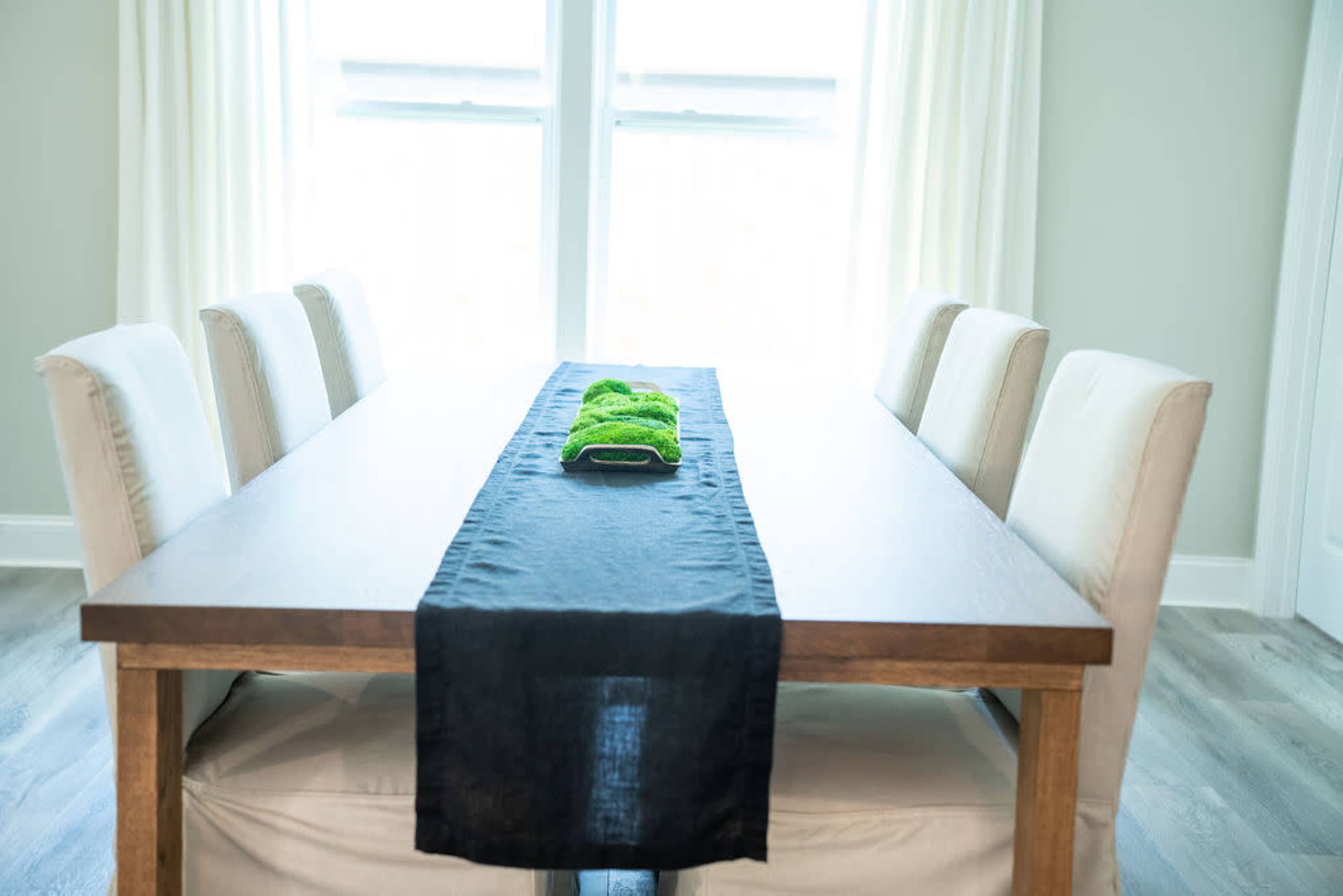 A wooden dining table with a black runner and a green centerpiece is surrounded by six upholstered chairs in a bright, airy room.