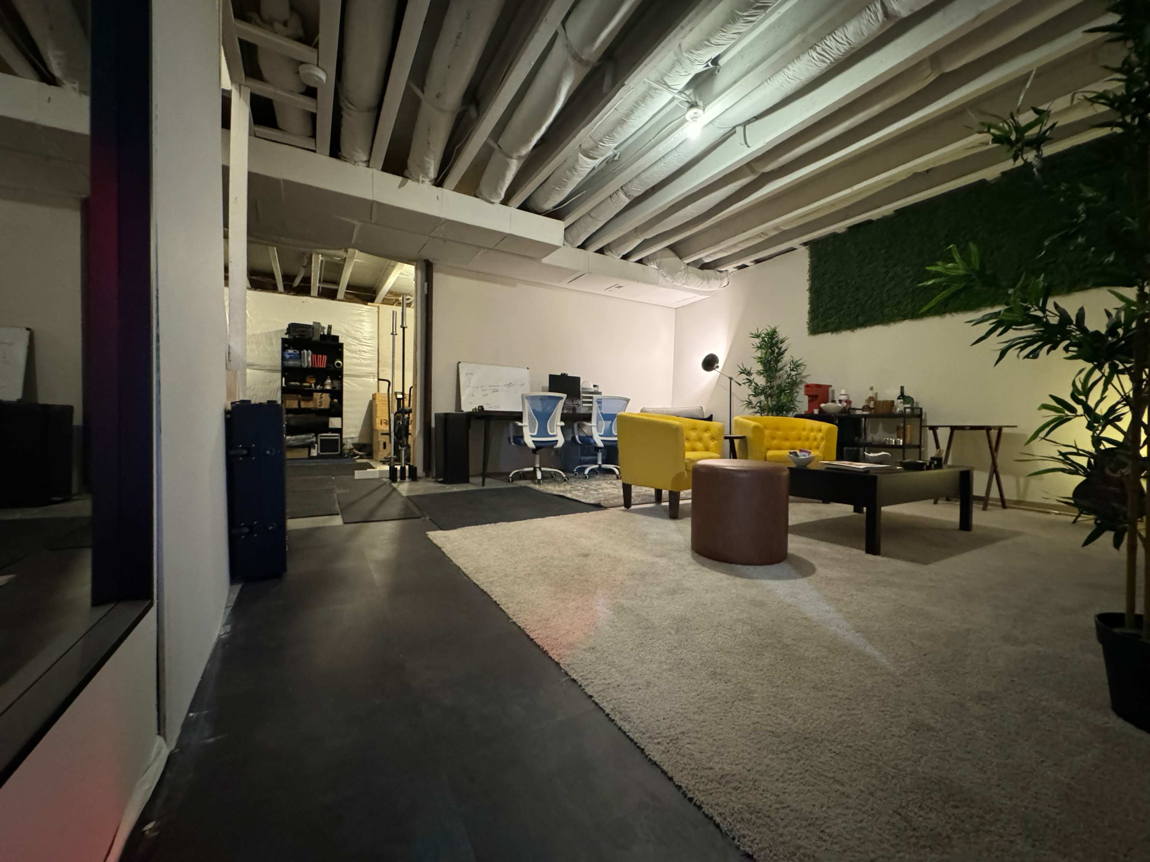 The image depicts a modern workspace featuring two blue chairs at a desk, a yellow couch, a round brown table, and green plants, all situated in a room with exposed ceiling beams and a carpeted floor.