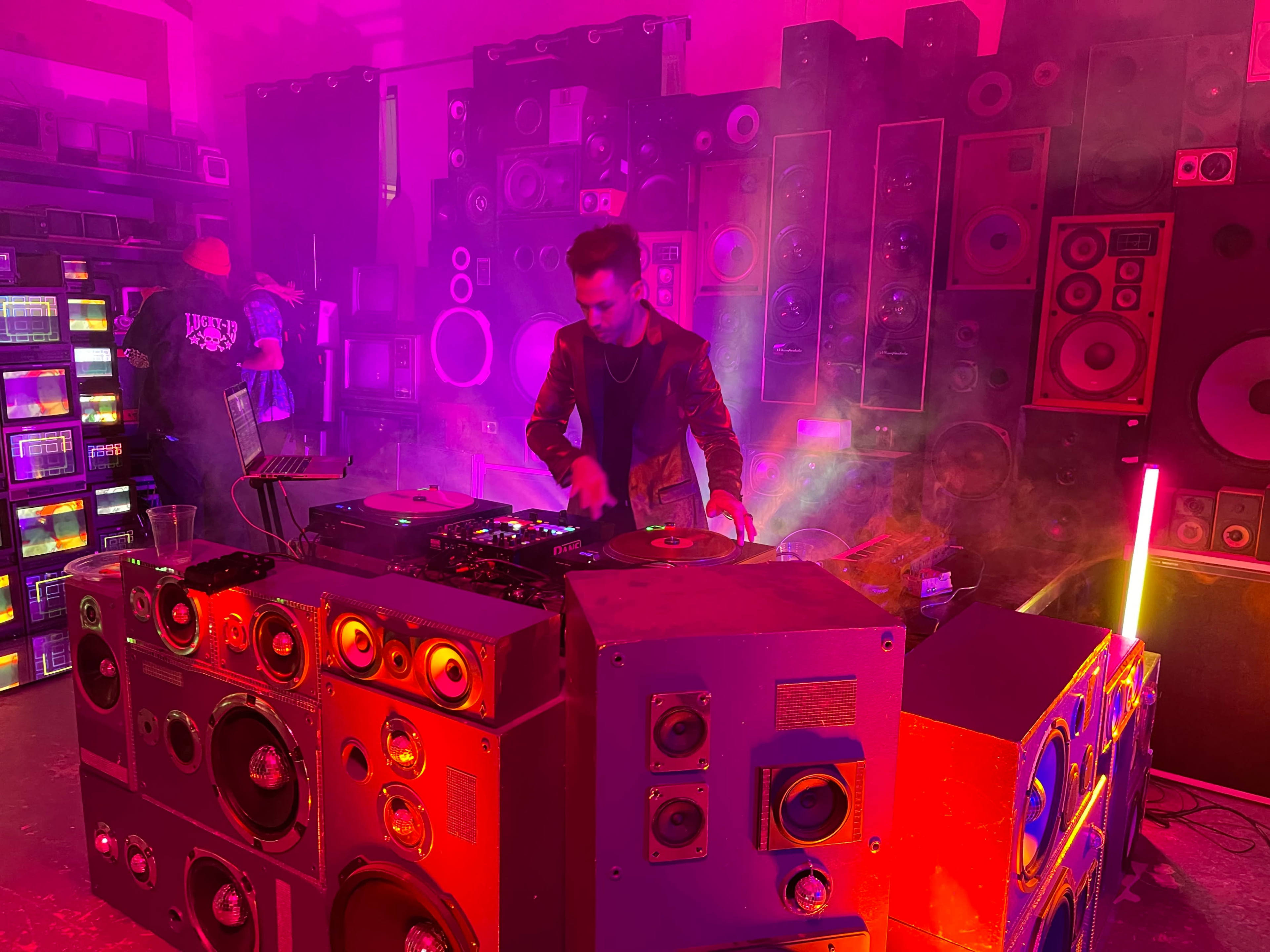A DJ performs in a room filled with colorful speakers and lights, surrounded by a mist of smoke.