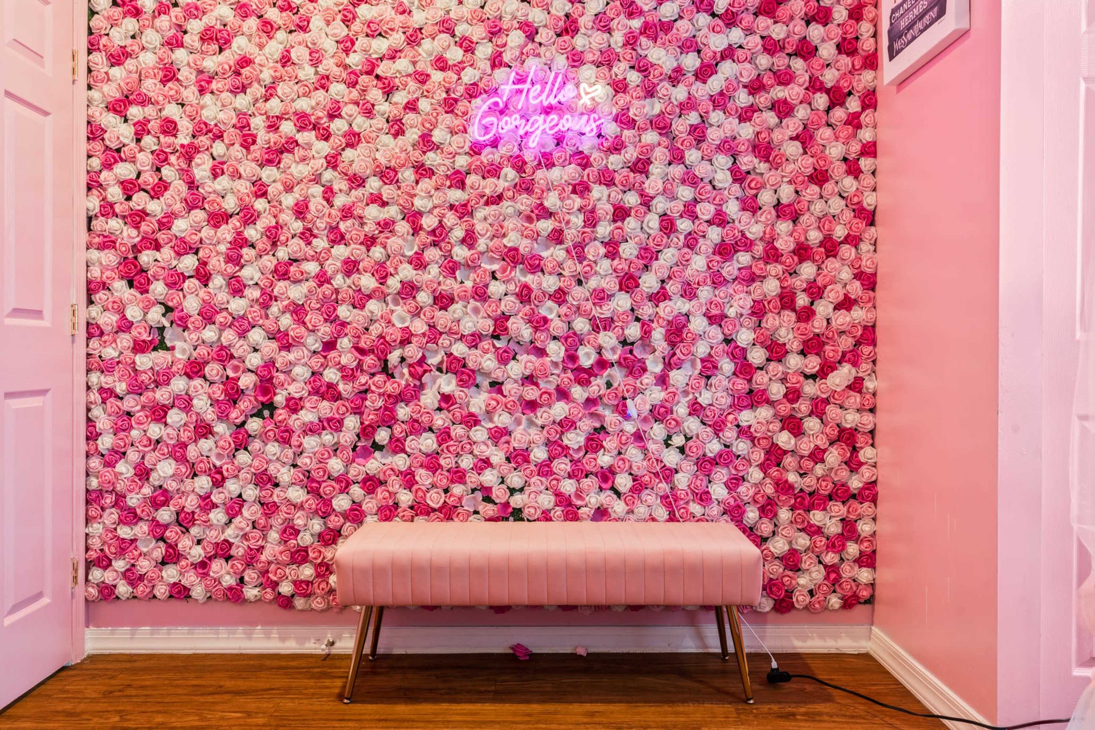A wall covered in faux roses in various shades of pink, with a neon sign that reads "Hello Gorgeous" and a pink bench in front of it.