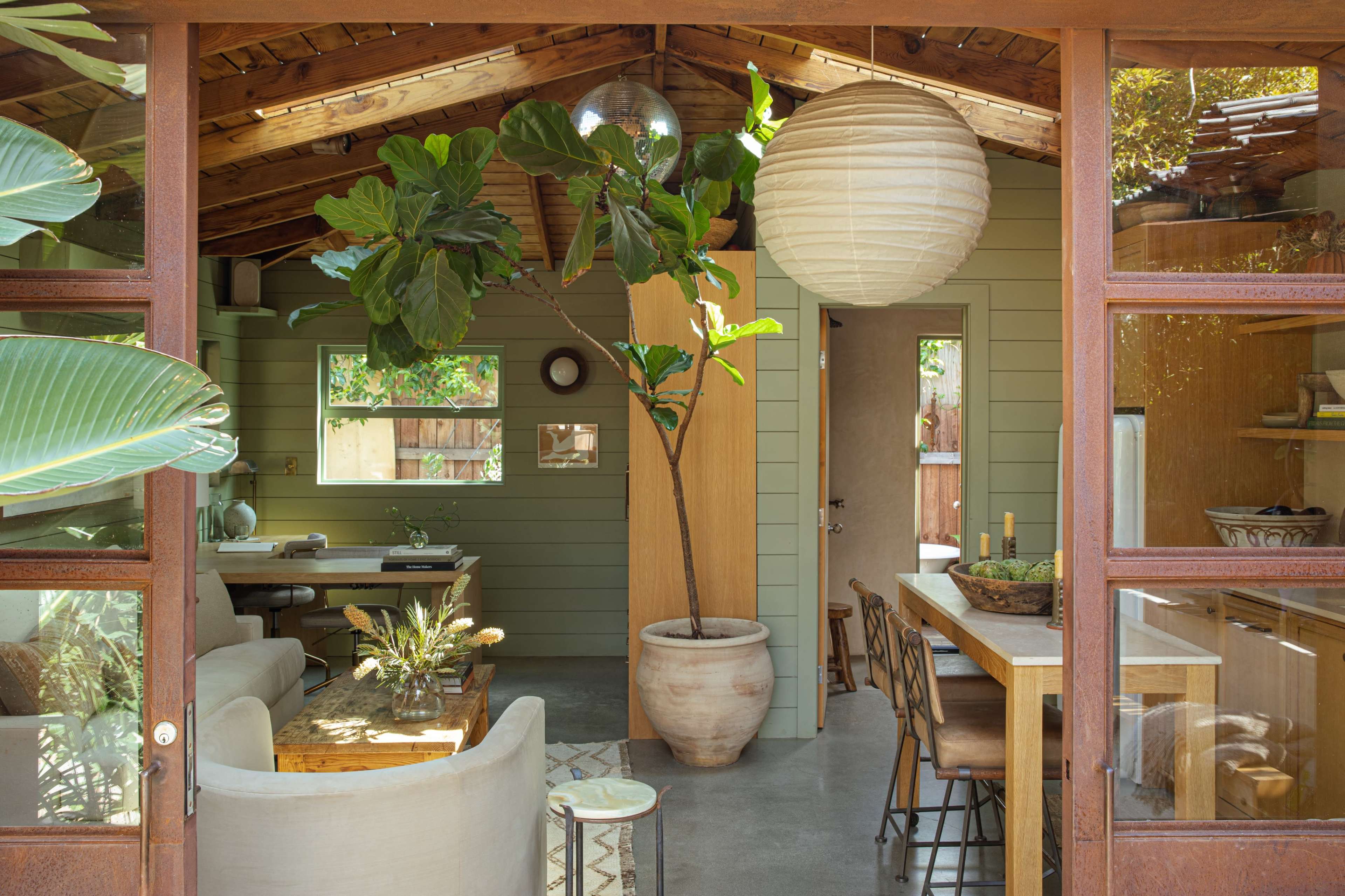 The image shows a modern, open-concept living space with a wooden ceiling, large windows, a dining area, and a potted plant.