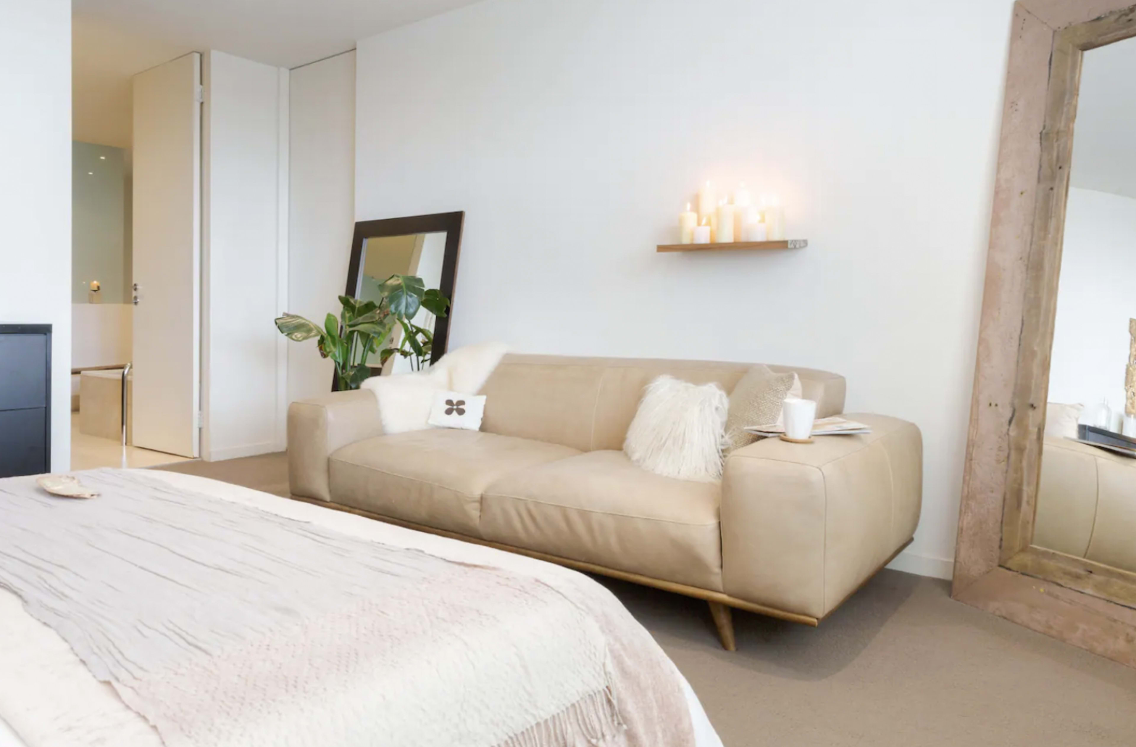 A light-colored leather sofa is positioned next to a large mirror and a small indoor plant, with soft decor elements on a nearby shelf.
