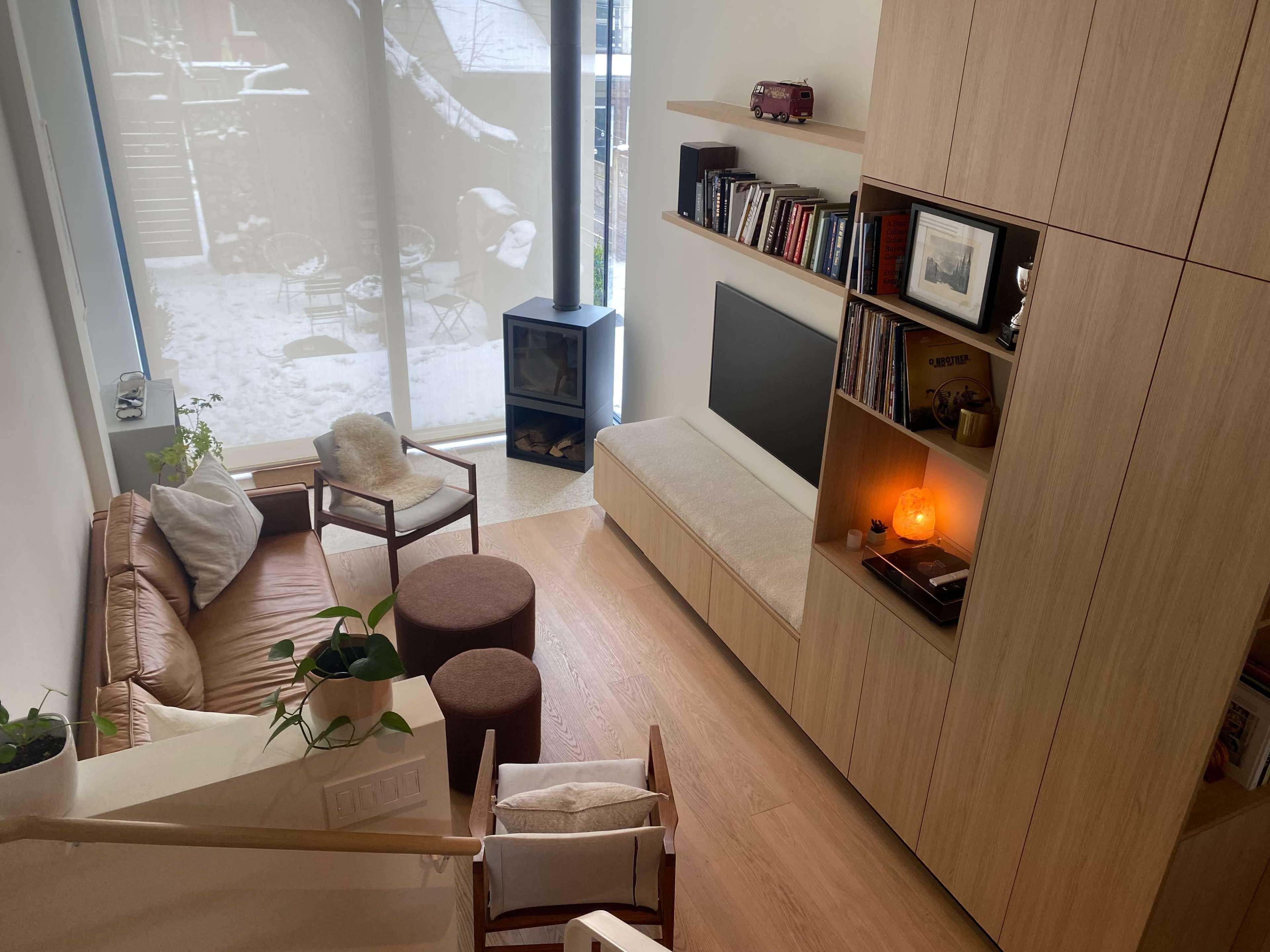 A cozy living space featuring a brown leather sofa, a wooden bookshelf with decor, a television, and a view of a winter scene through a large window.