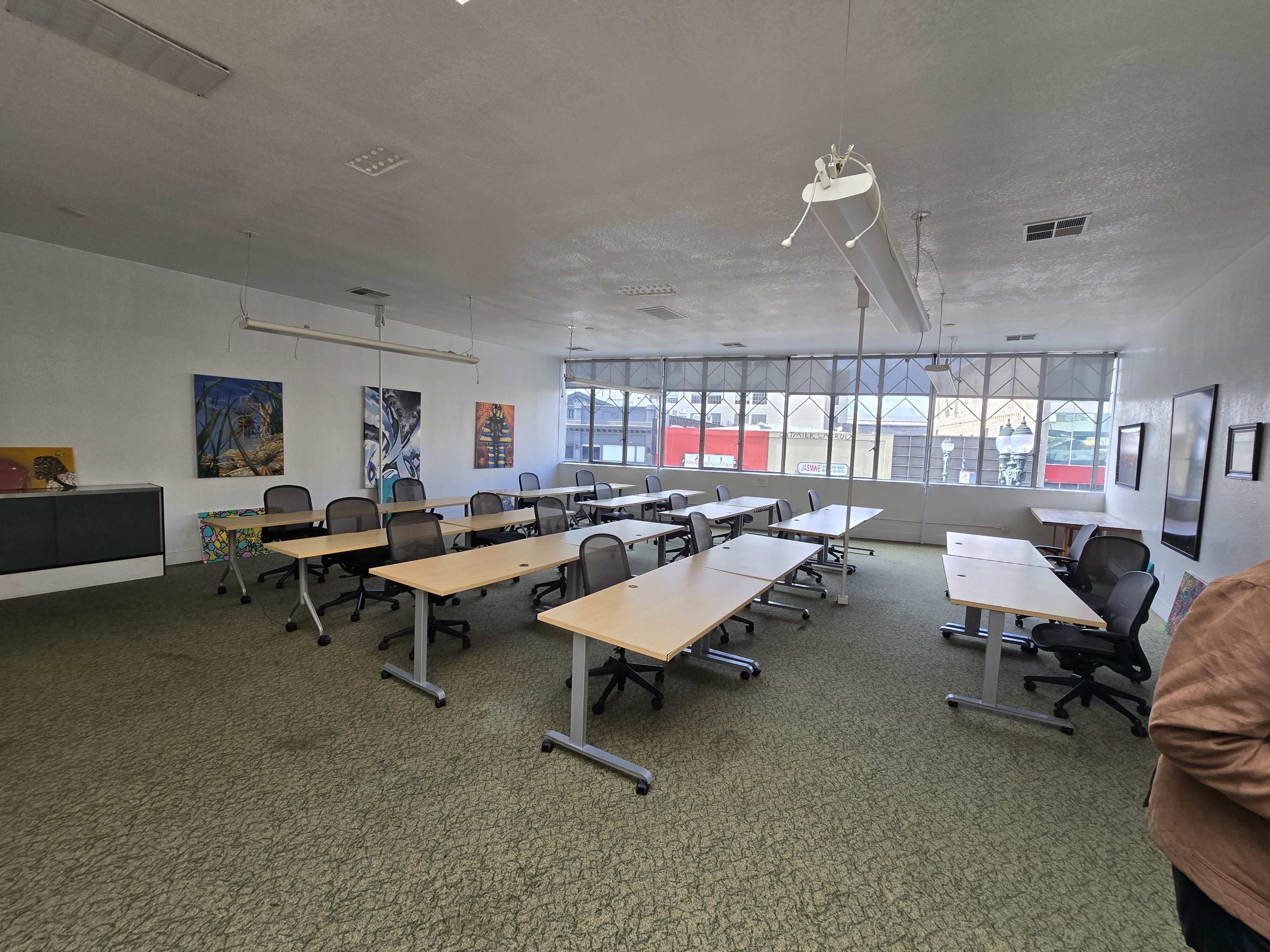 The image shows a spacious, empty classroom or meeting room with multiple desks and chairs arranged in rows and large windows along one wall.