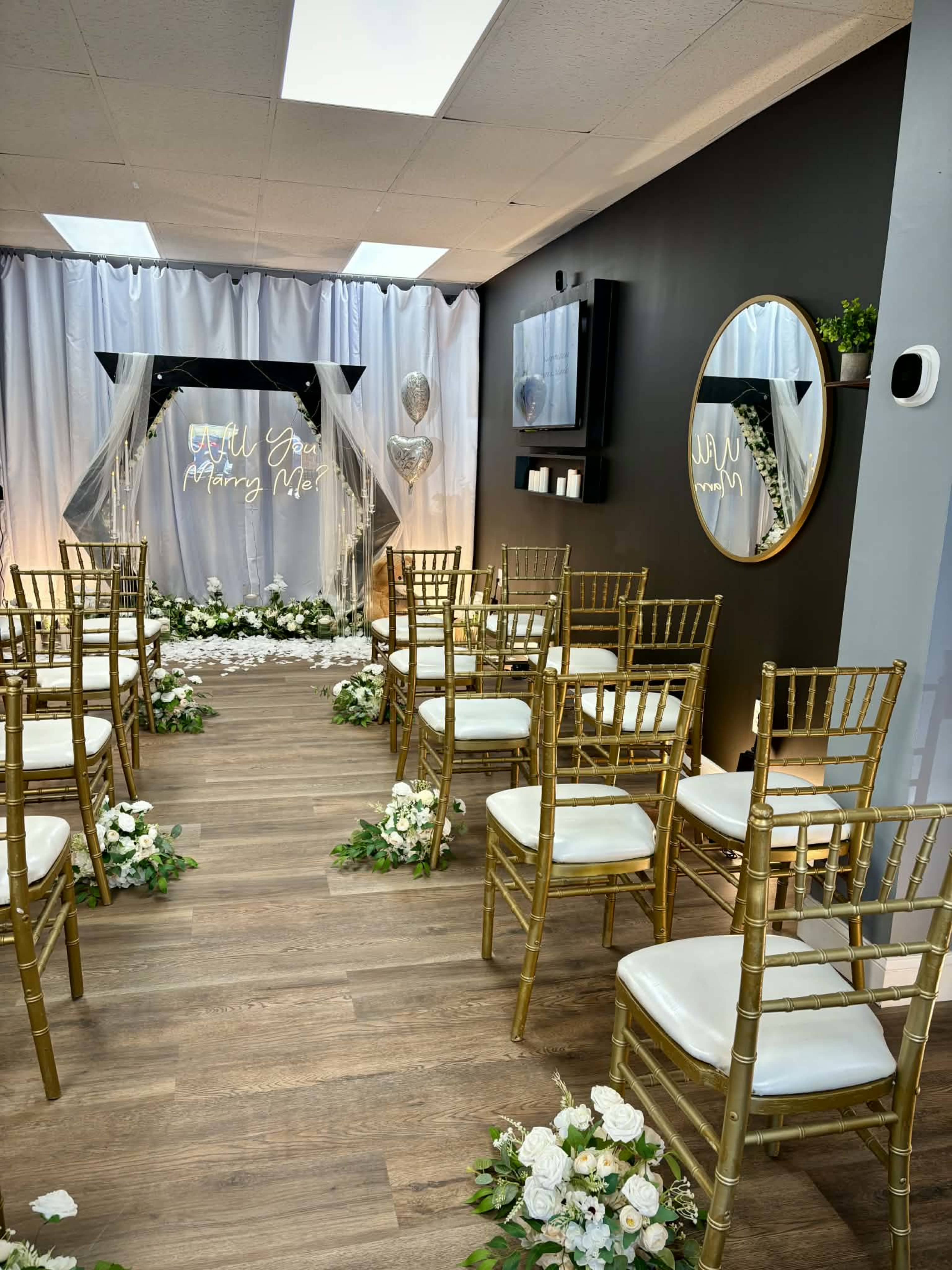 The image shows a wedding proposal setup with gold chairs arranged in front of a decorated arch, surrounded by floral arrangements and draped fabric in a well-lit indoor space.