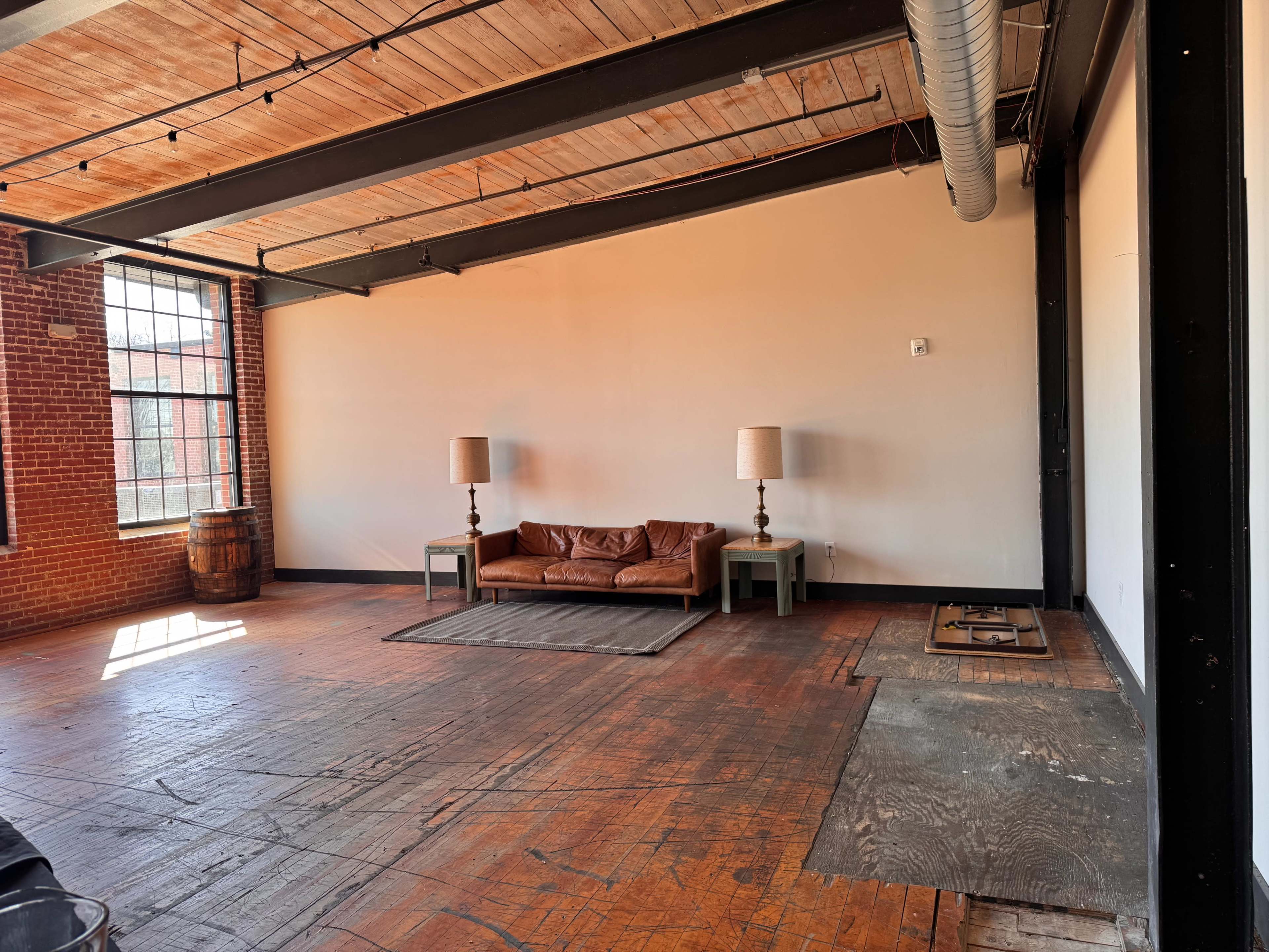 The image shows a spacious, industrial-style room featuring a brown leather couch, two floor lamps on either side, and a barrel in the corner, with large windows letting in natural light.