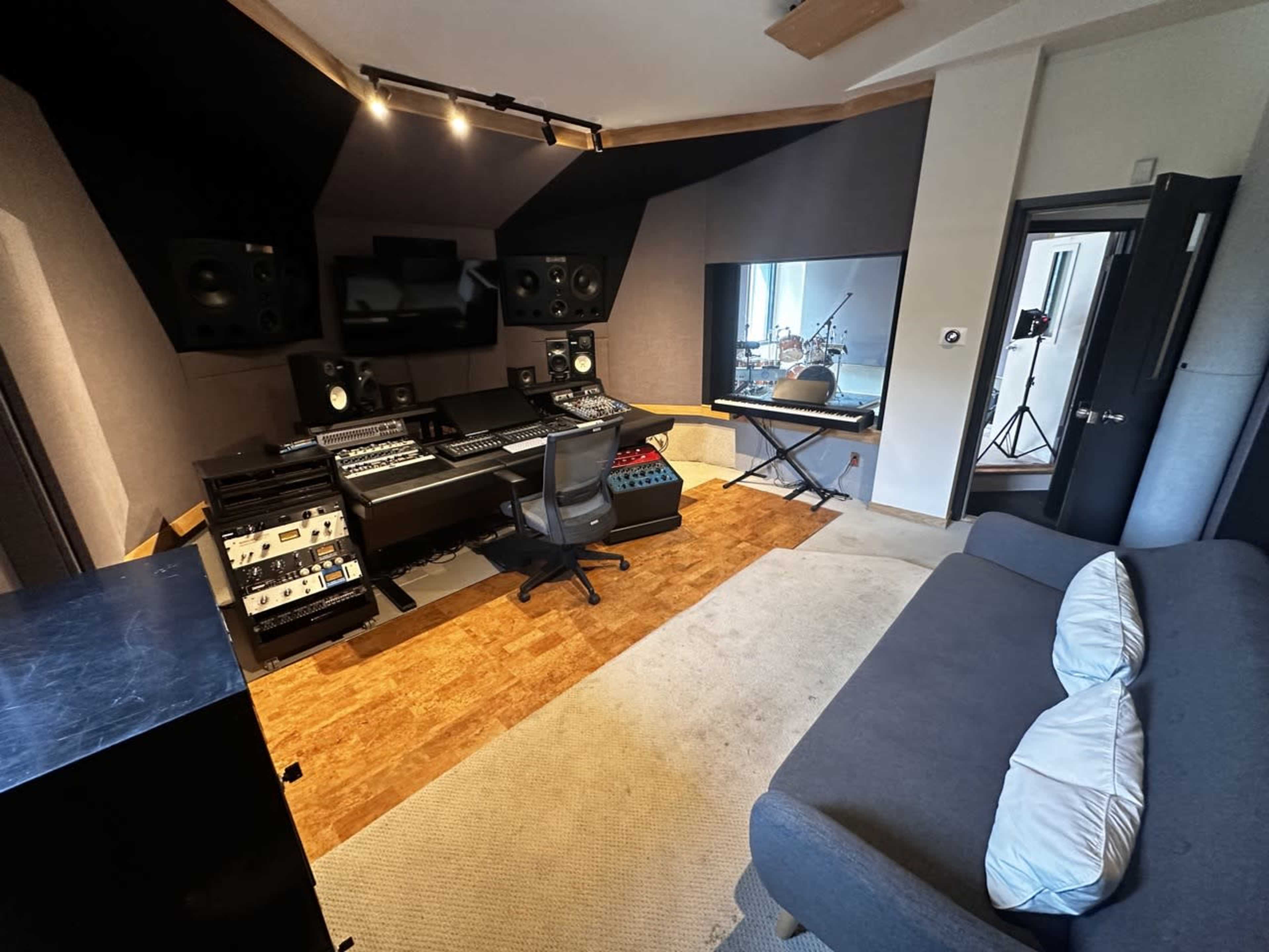 The image shows a modern recording studio with sound equipment, a mixing console, monitors, and a seating area.