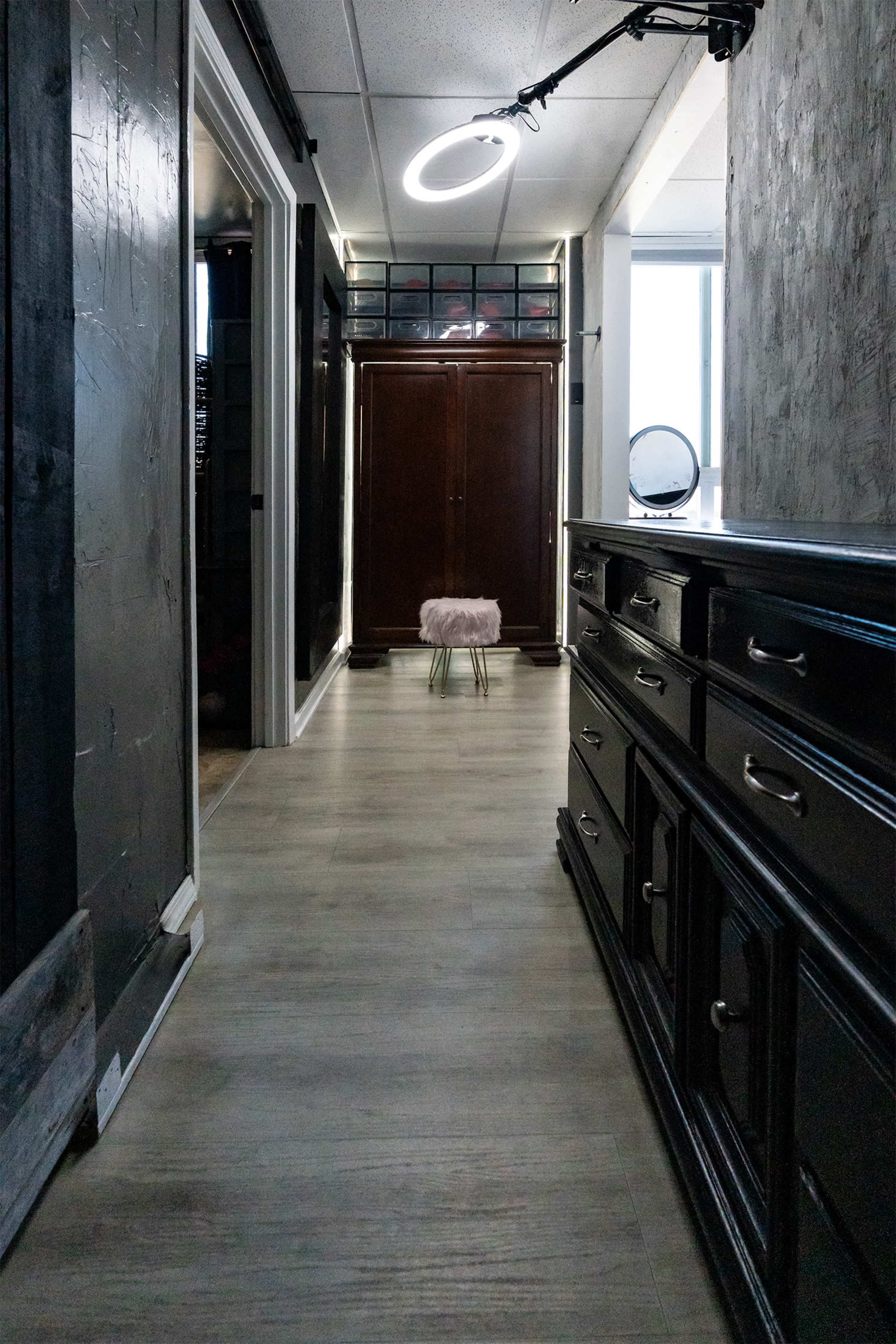 A narrow hallway features a dark wood dresser, a plush stool at the end, and soft lighting from a circular fixture above.