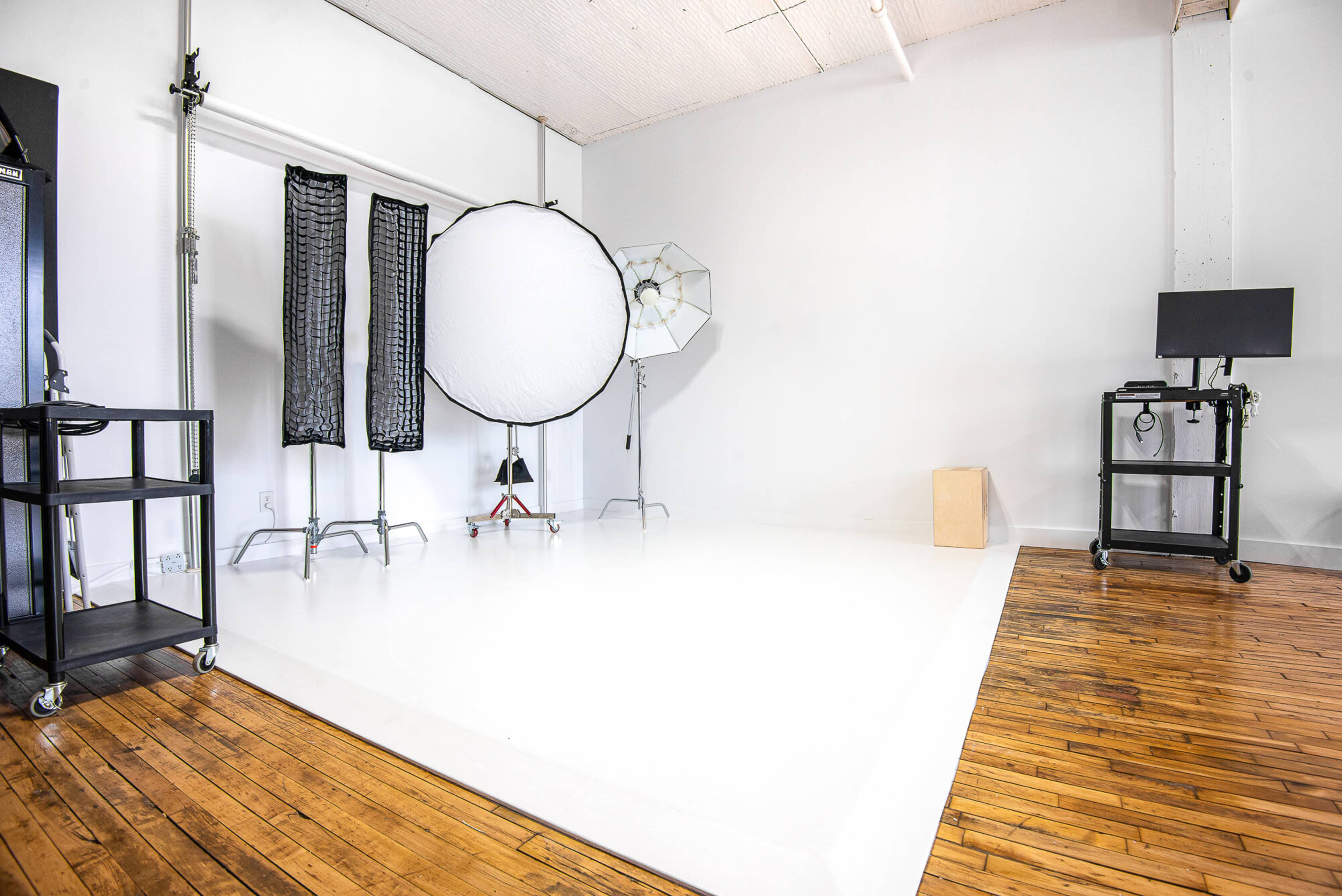 The image shows a photography studio with white walls, a polished wooden floor, and various lighting equipment set up, including softboxes and backdrops.