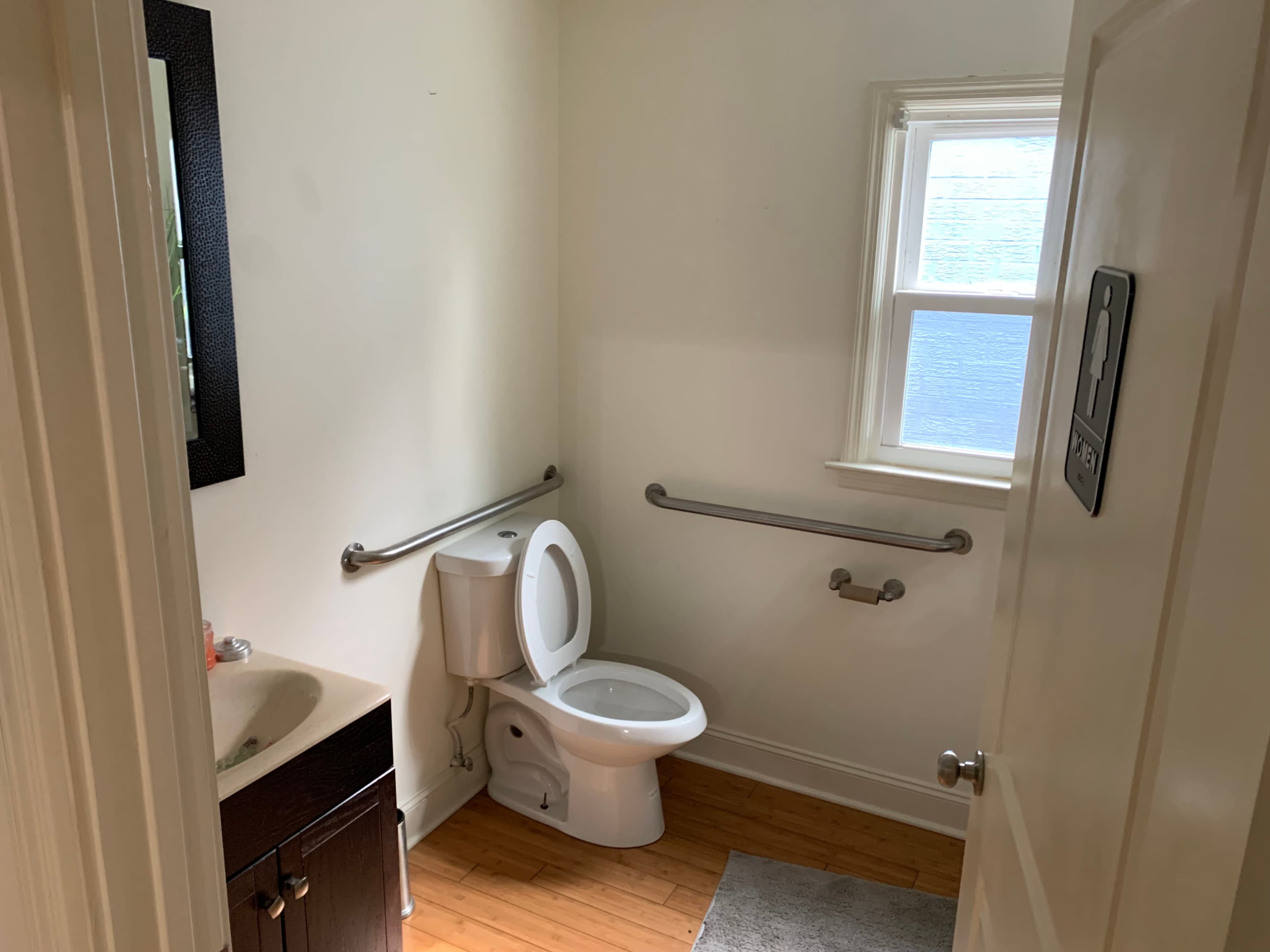 The image shows a small bathroom with a toilet, a sink, and grab bars on the walls, illuminated by natural light from a nearby window.