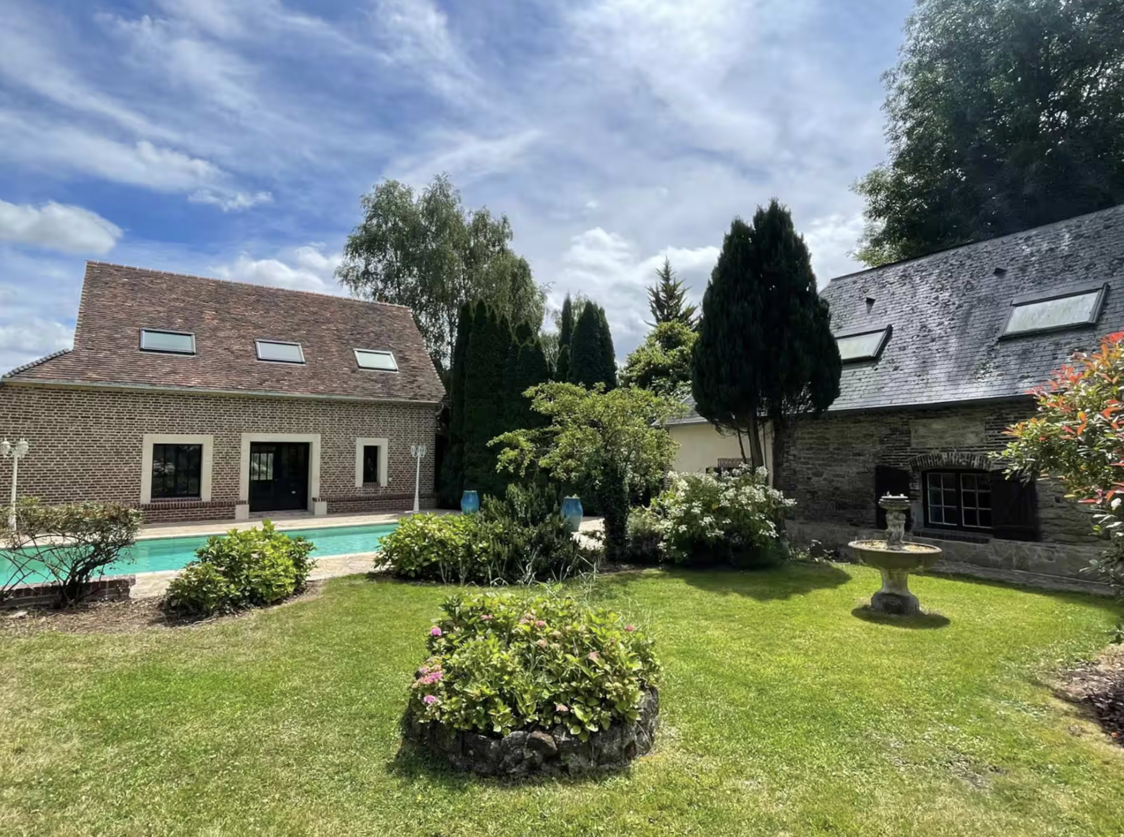 The image shows a landscaped garden with a swimming pool and two houses, one modern and one with stone walls, under a partly cloudy sky.