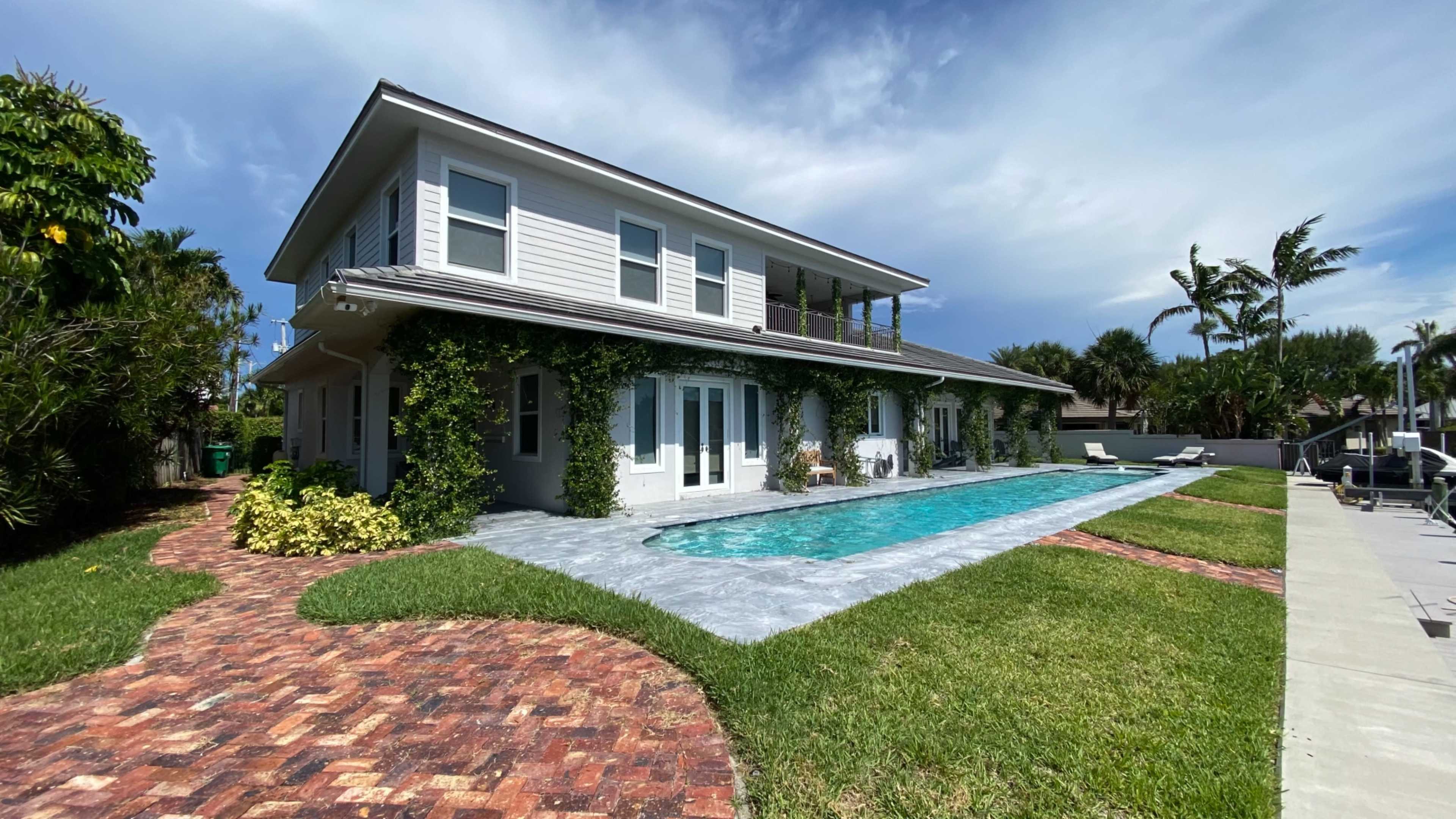 A two-story house with a pool and landscaped gardens is outlined by brick pathways and surrounded by palm trees.