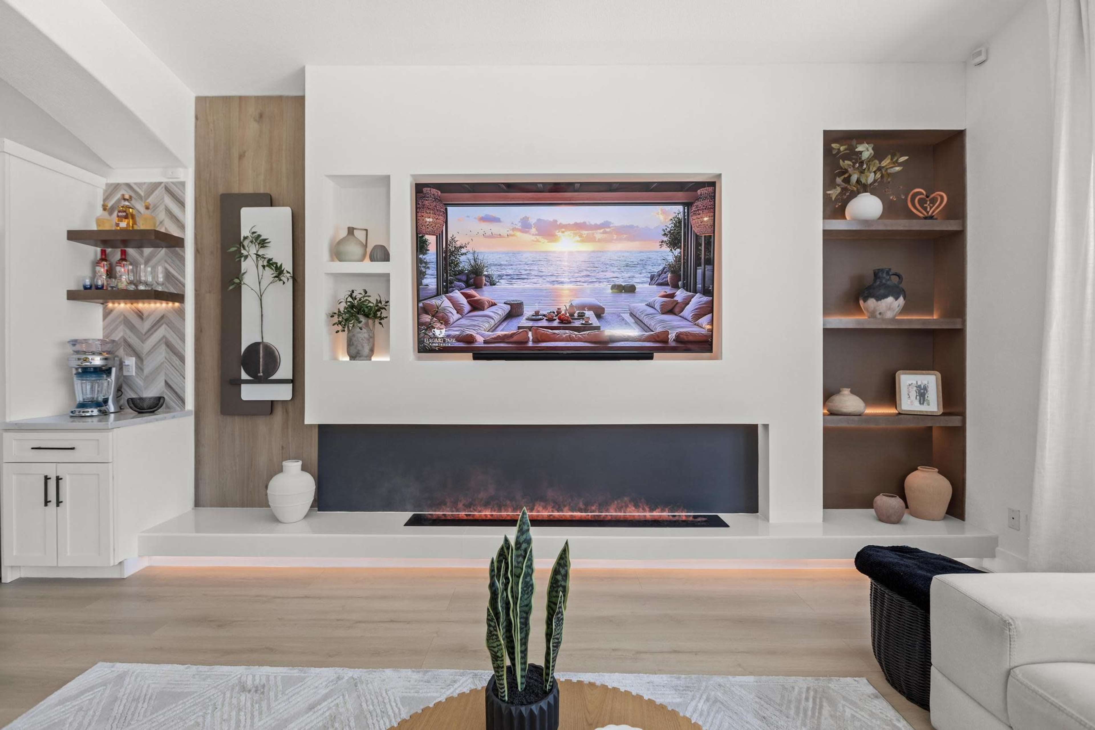 A modern living room features a sleek entertainment center with a large TV displaying a sunset over a beach, complemented by a minimalist fireplace and decorative shelves.