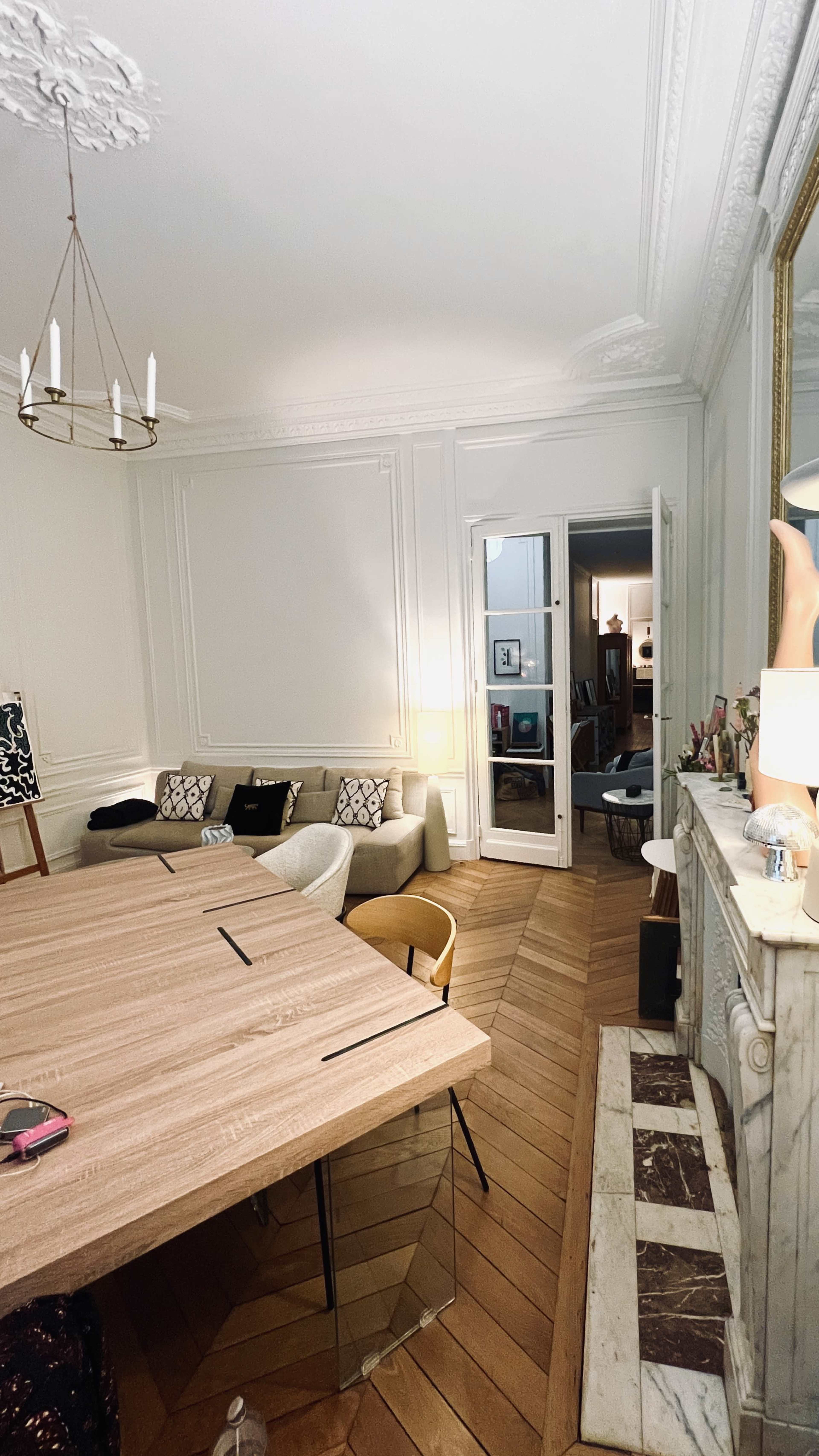 The image showcases a well-lit room with wooden flooring, a large table, and a modern fireplace, featuring seating arrangements and a doorway leading to another space.