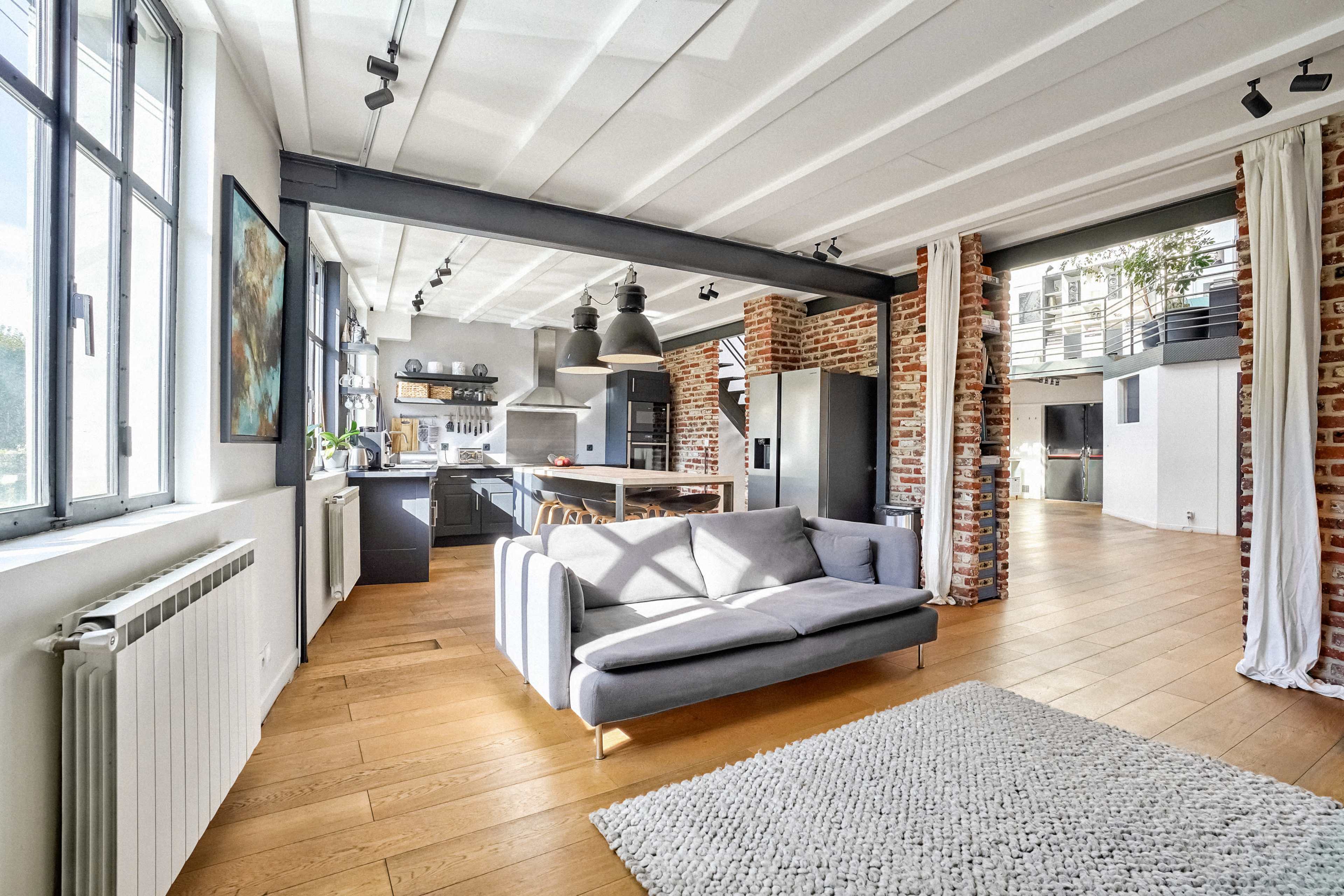 The image shows a modern, open-concept living space featuring a gray sofa, exposed brick walls, and a large kitchen with industrial-style lighting.
