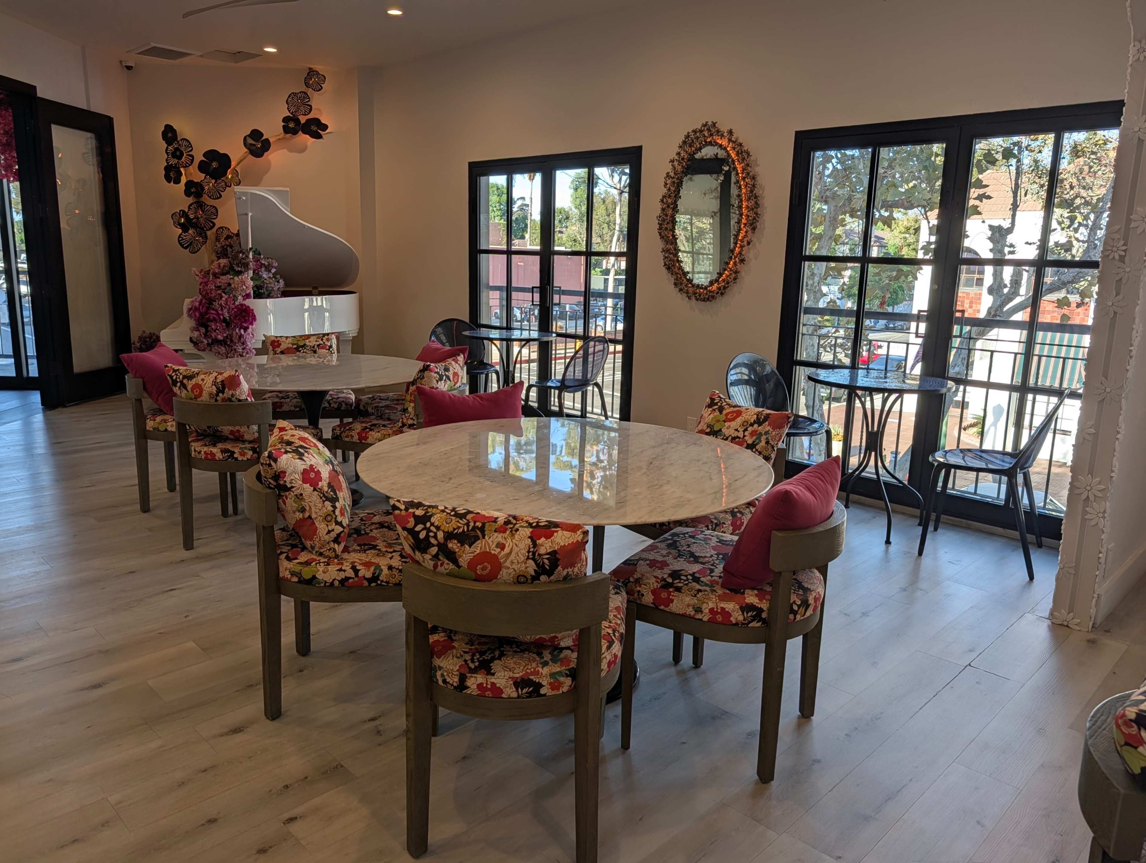 The image shows a bright café interior with round marble tables surrounded by upholstered chairs featuring floral patterns, and windows allowing natural light to enter.