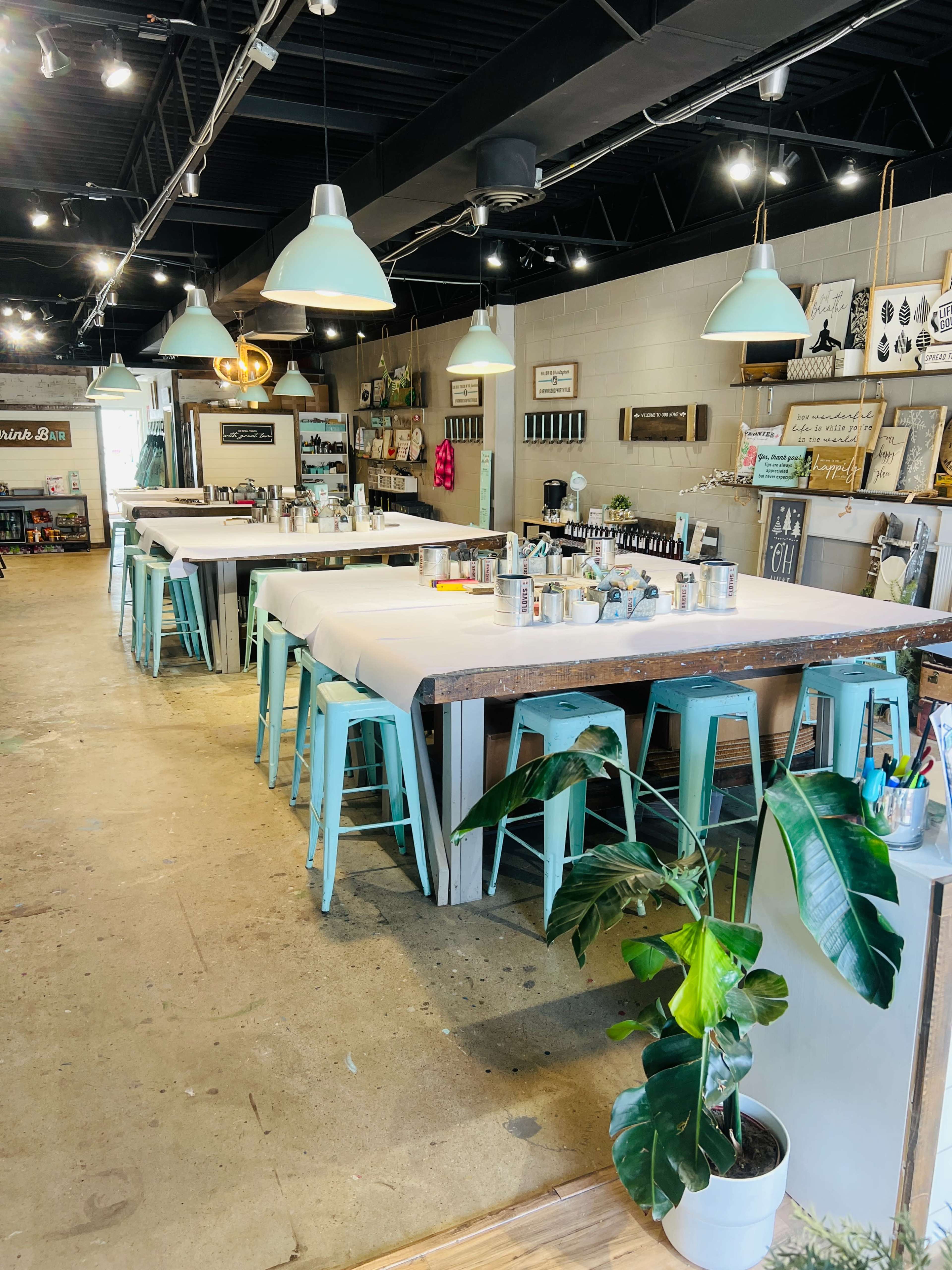 A spacious, well-lit creative studio with several large tables surrounded by turquoise chairs and various art supplies displayed around the room.