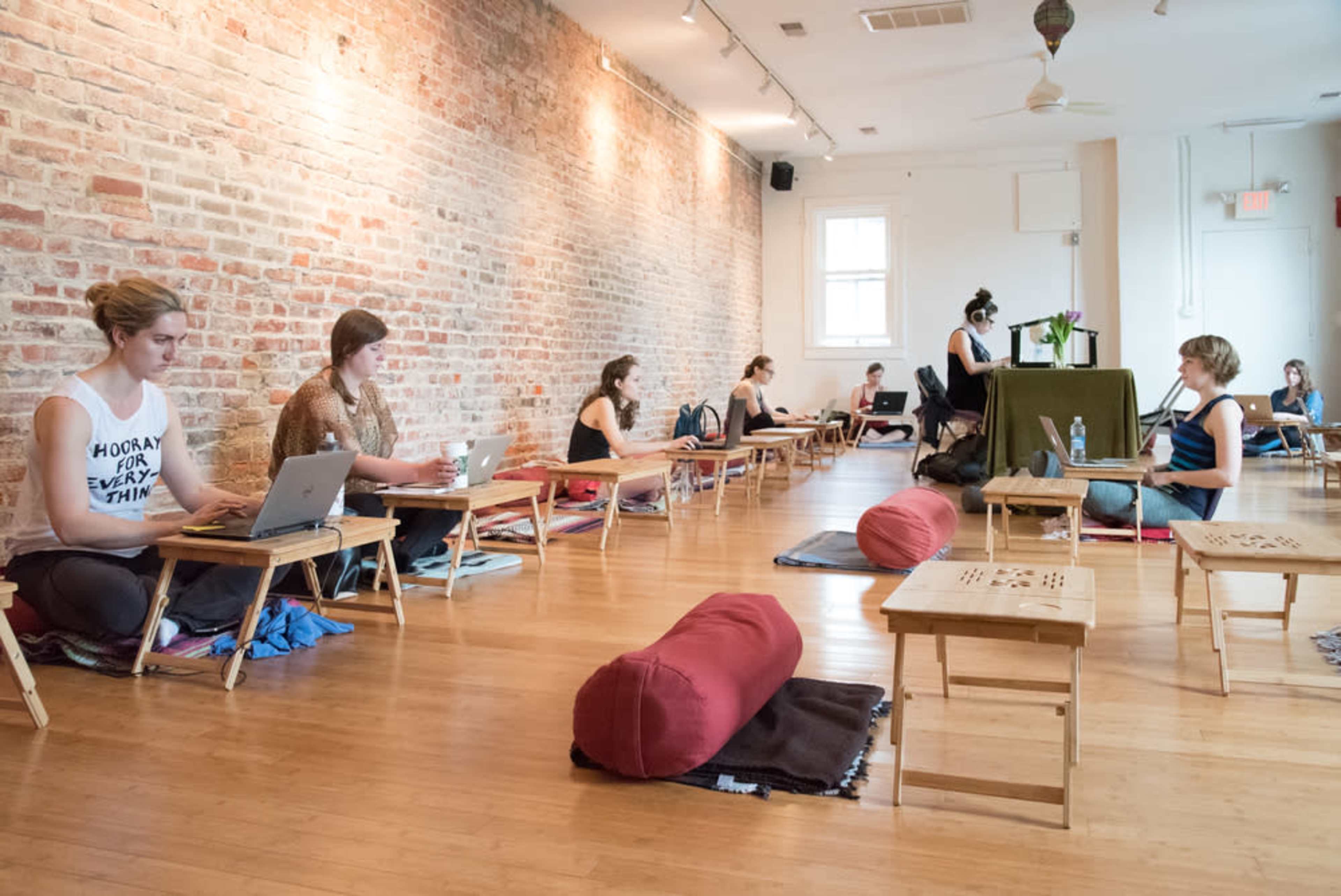 A group of people is working on laptops in a spacious, well-lit room with exposed brick walls and low wooden tables.