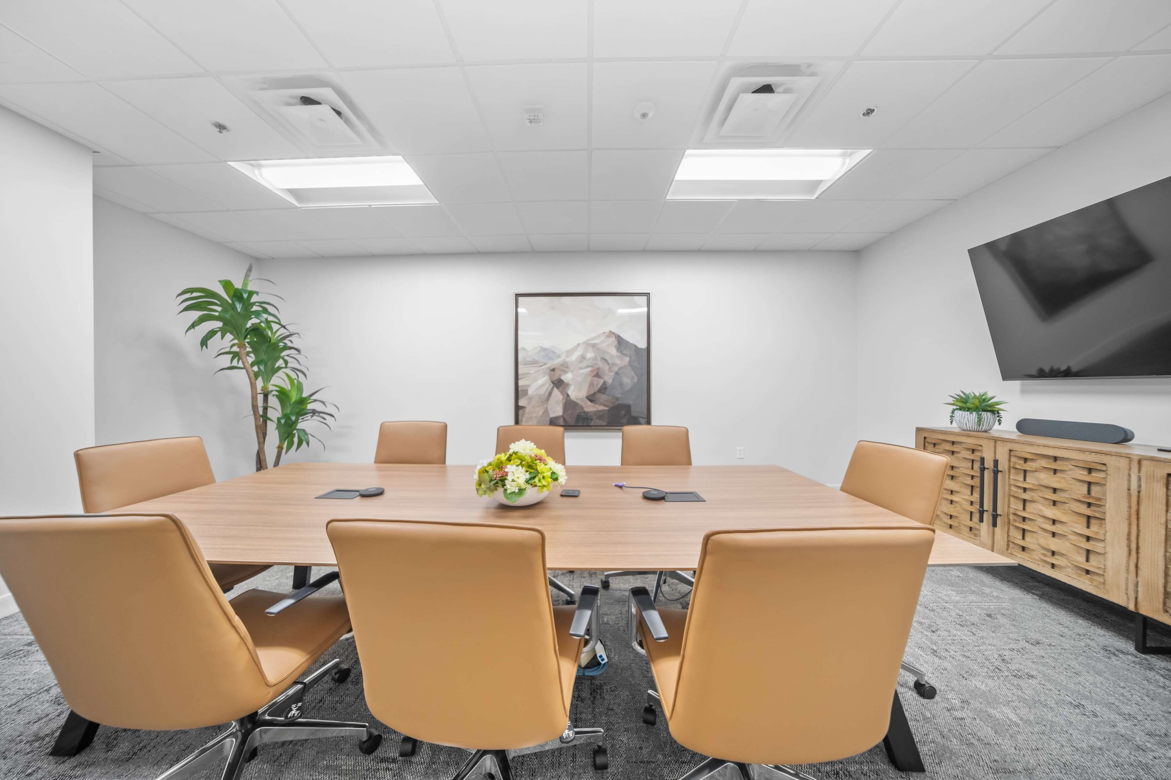 A modern conference room features a large wooden table surrounded by six orange chairs, with a plant, artwork, and a television on the walls.