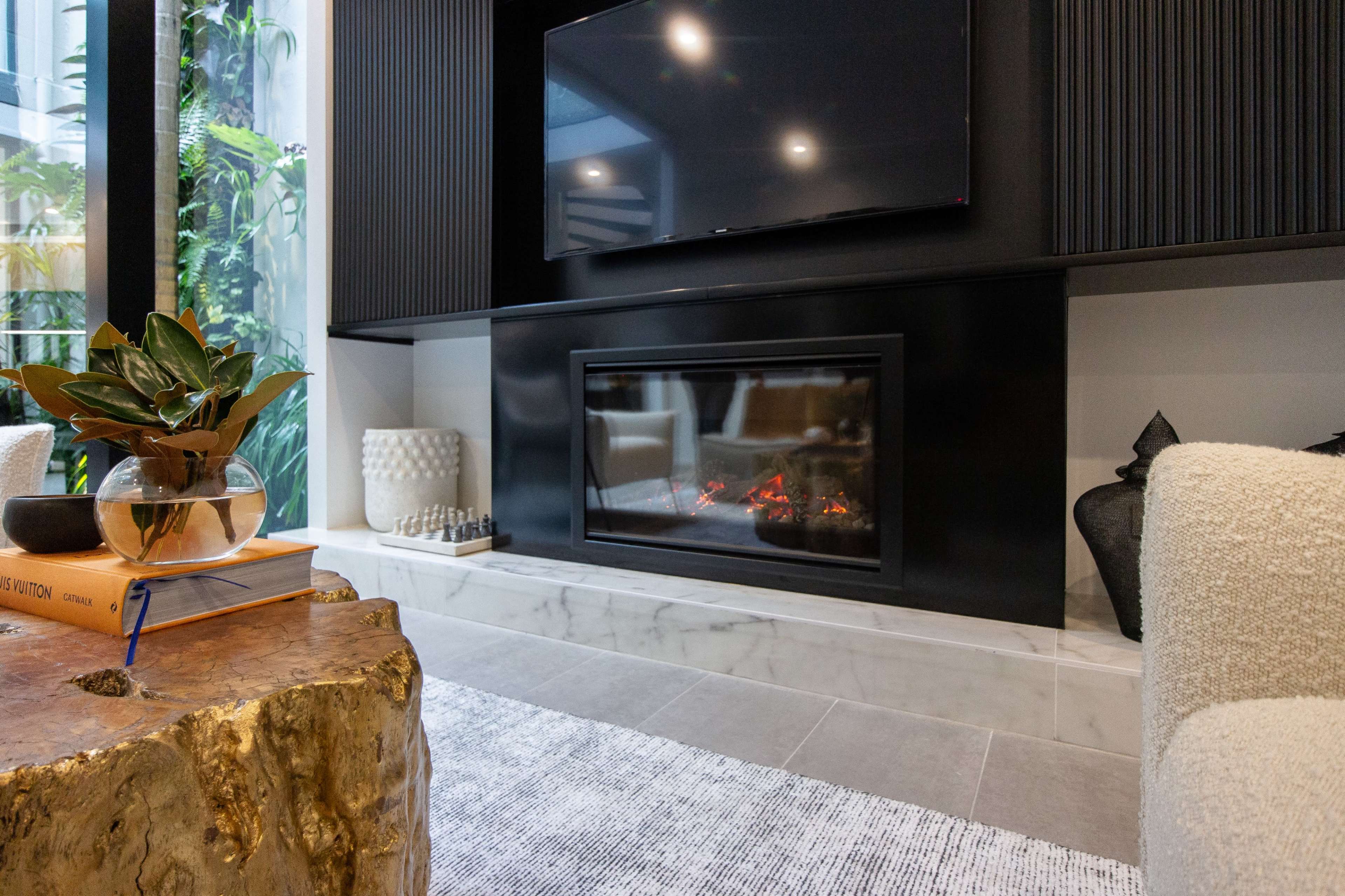 A living area with a modern black fireplace beneath a television, surrounded by plants and decorative elements on a marble hearth.