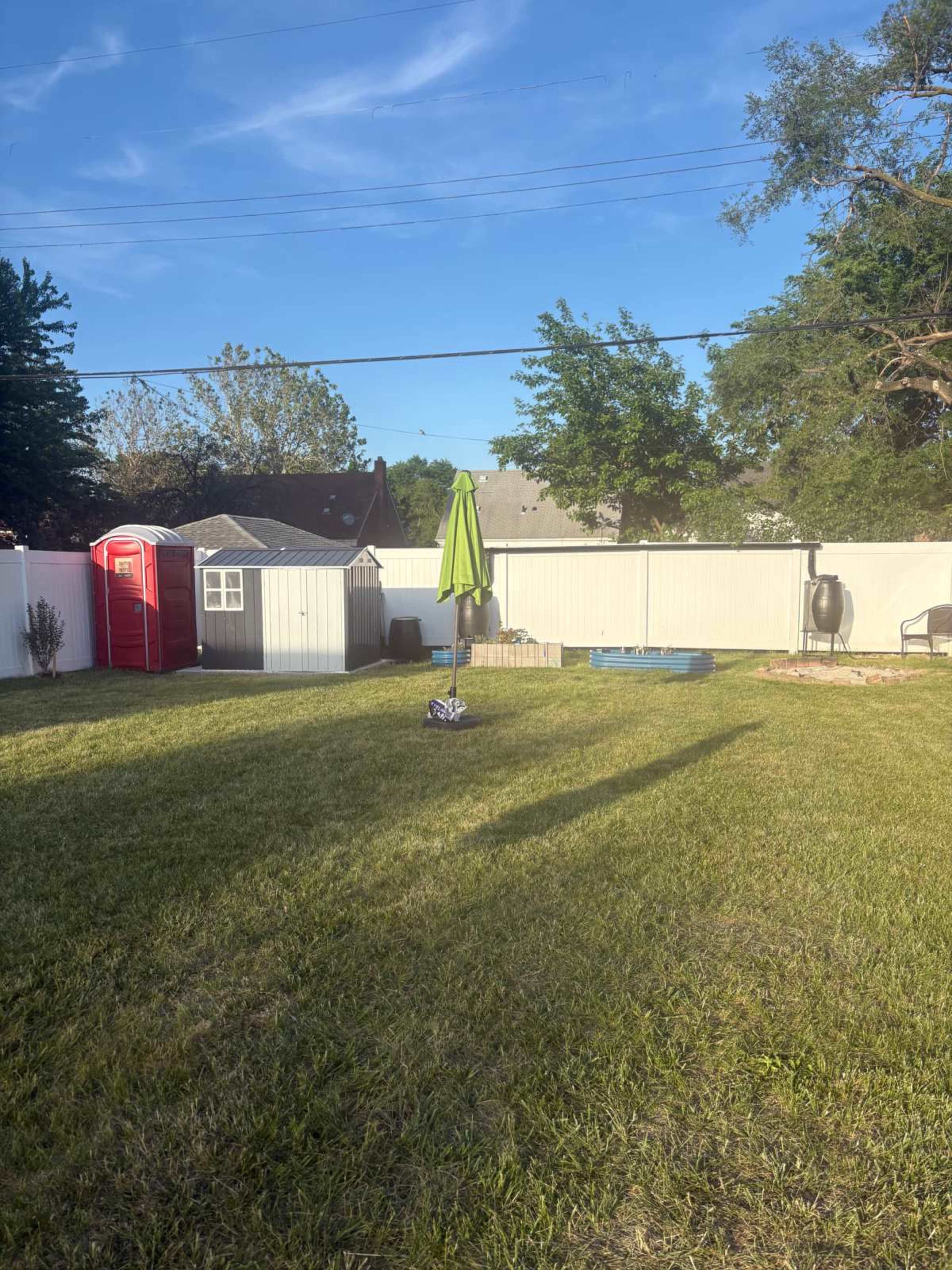 The image shows a spacious backyard with a green umbrella, a small shed, and a blue pool surrounded by a white fence.