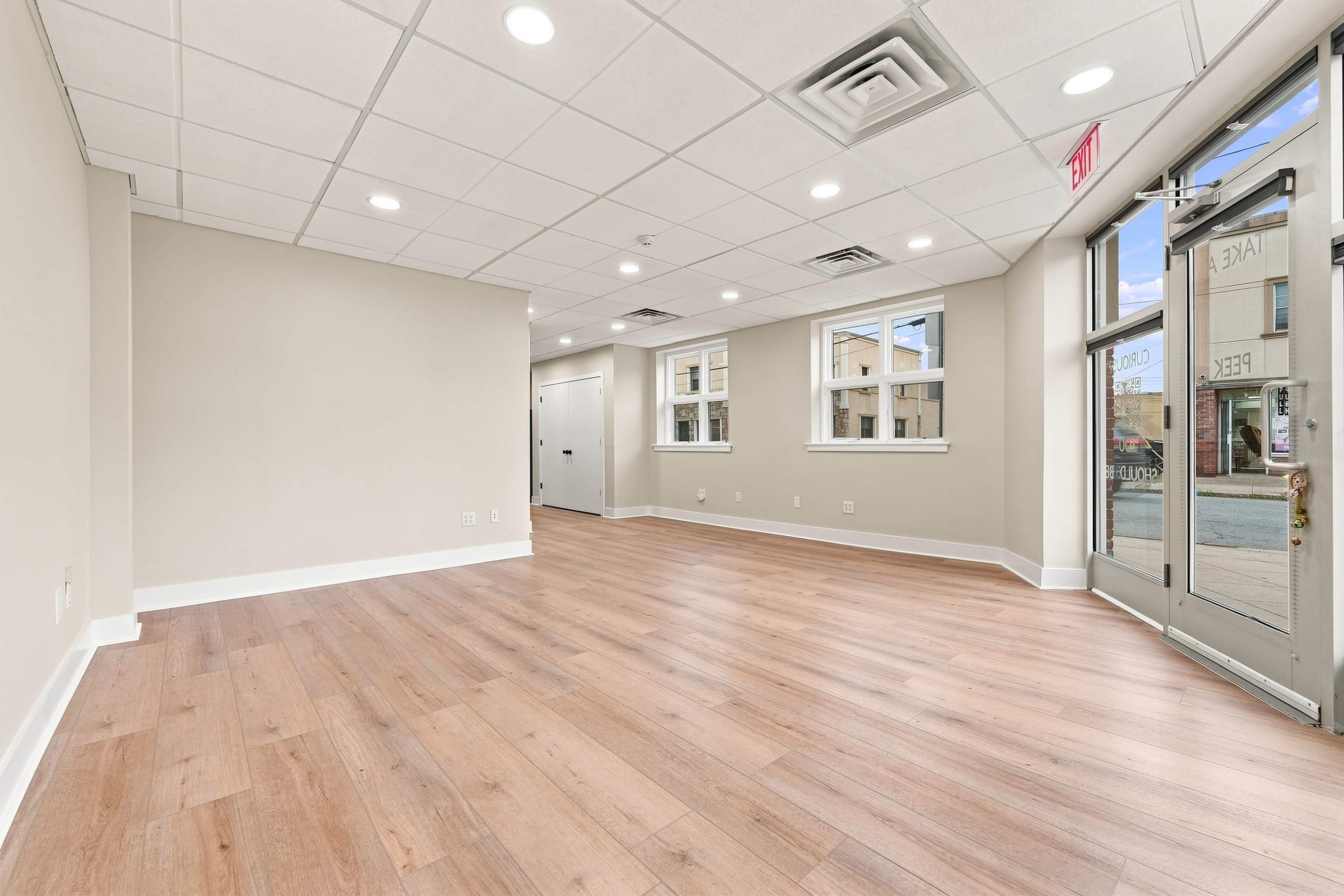 The image shows a vacant interior space with light-colored walls, a wooden floor, and large windows allowing natural light.