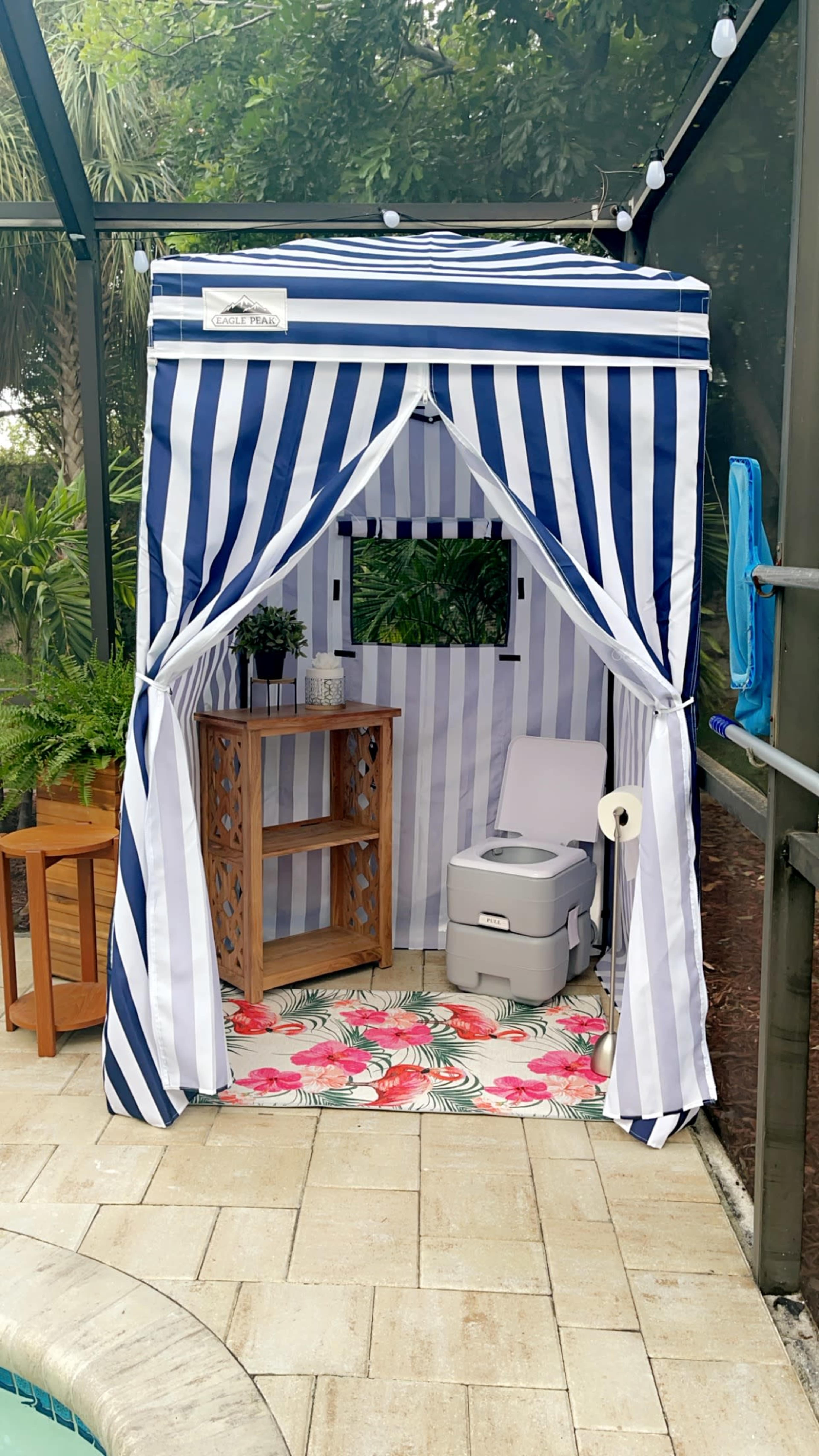 A striped privacy tent with a portable toilet inside is set up beside a pool, featuring a decorative mat and a small wooden table.