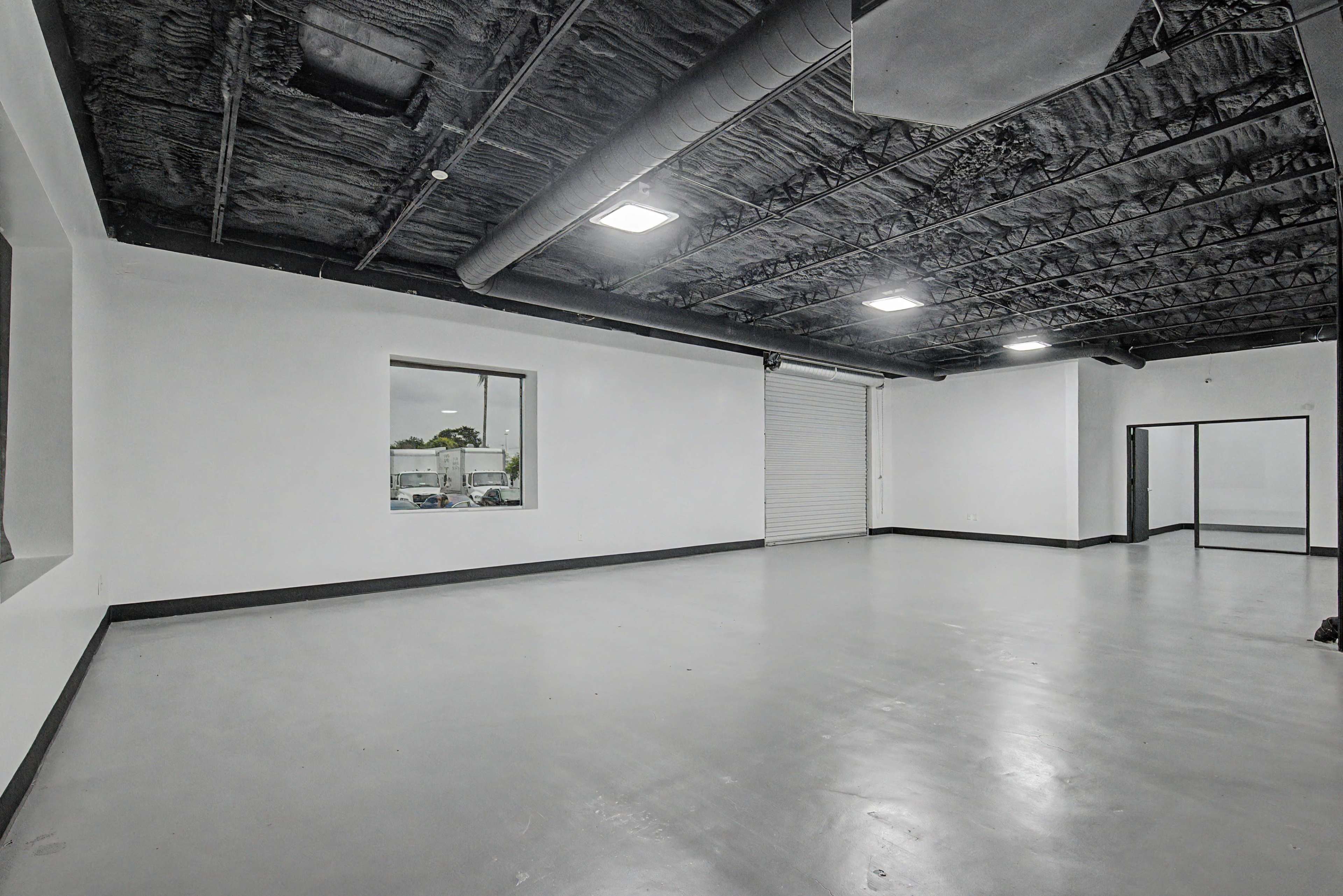 The image shows an empty room with white walls, a concrete floor, and a black ceiling, featuring windows and a roll-up door.