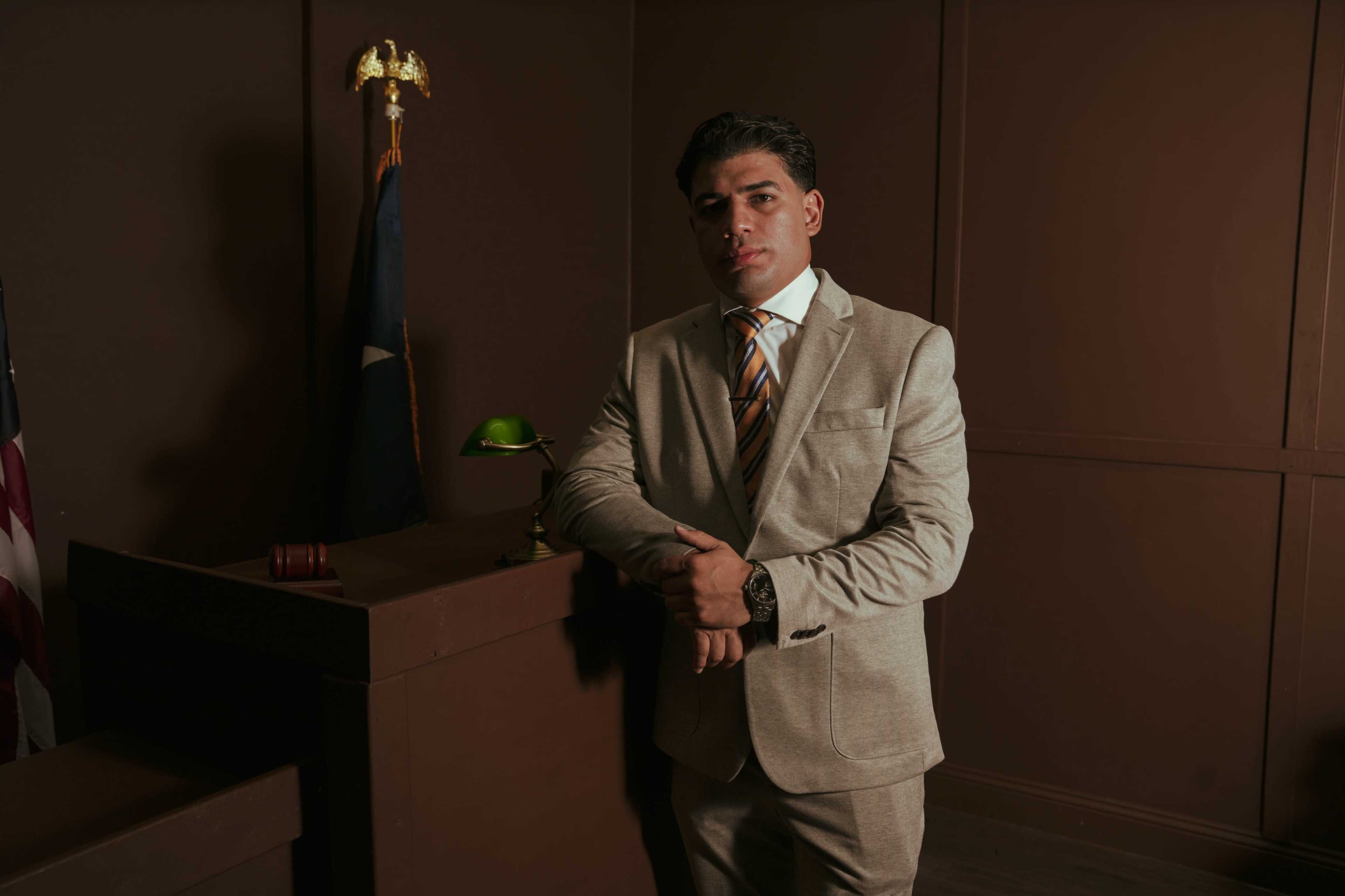 A man in a beige suit stands confidently with his arms crossed in a dimly lit courtroom, featuring a gavel and a green desk lamp.