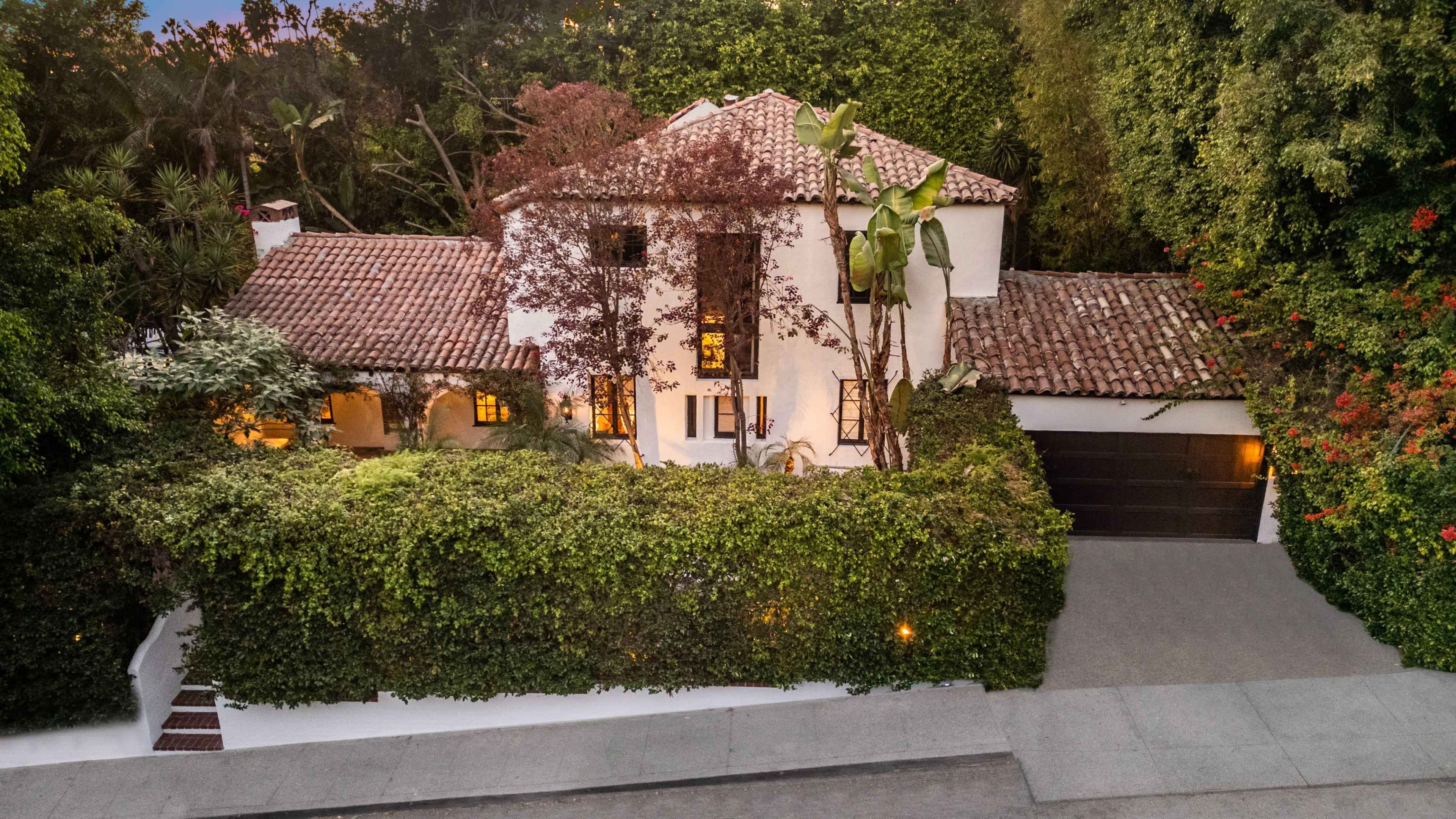 A white stucco house with a red-tiled roof is surrounded by lush greenery and features a garage on the right side.