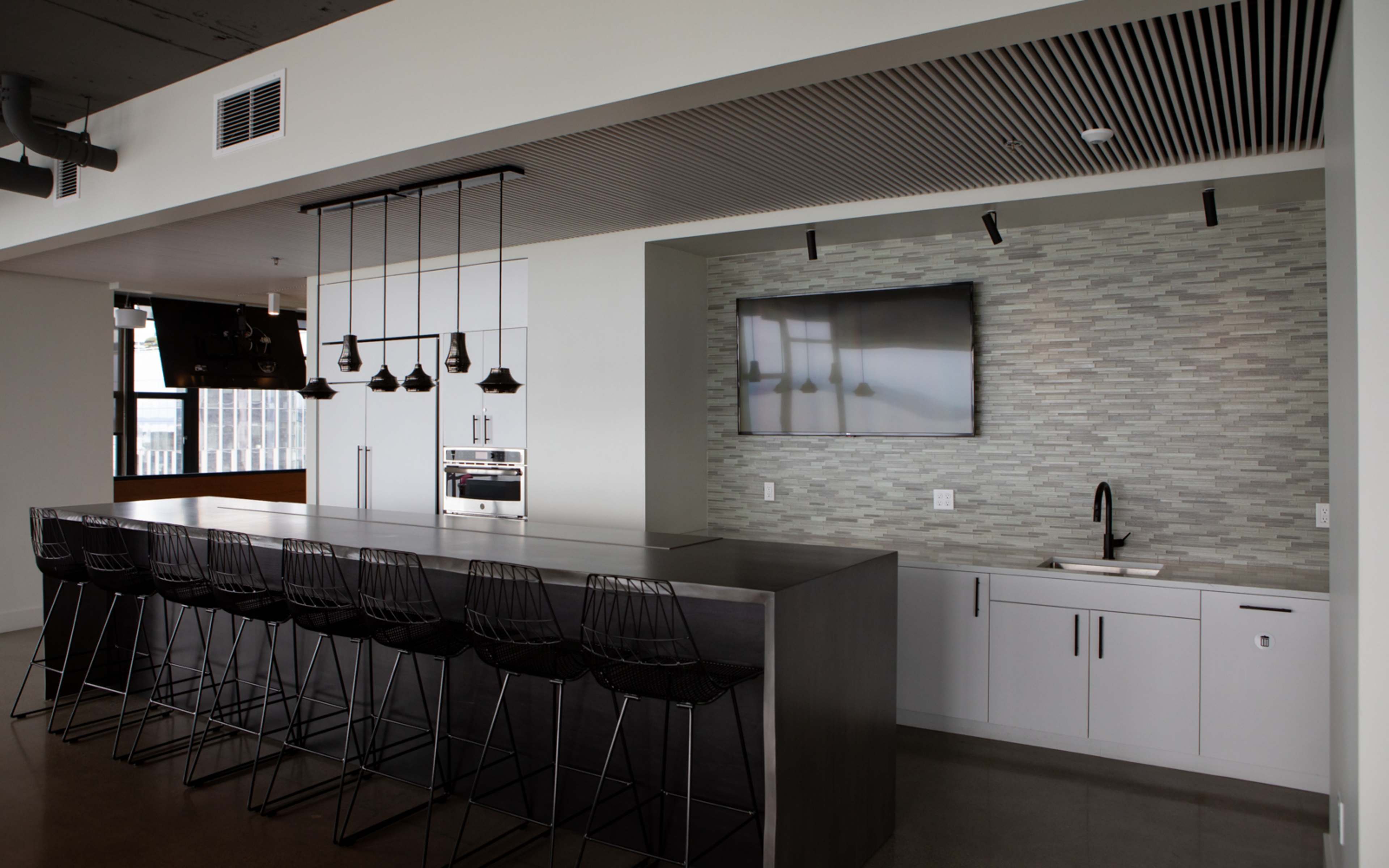 A modern kitchen with a long gray countertop, black bar stools, and a wall-mounted television above a sleek backsplash.