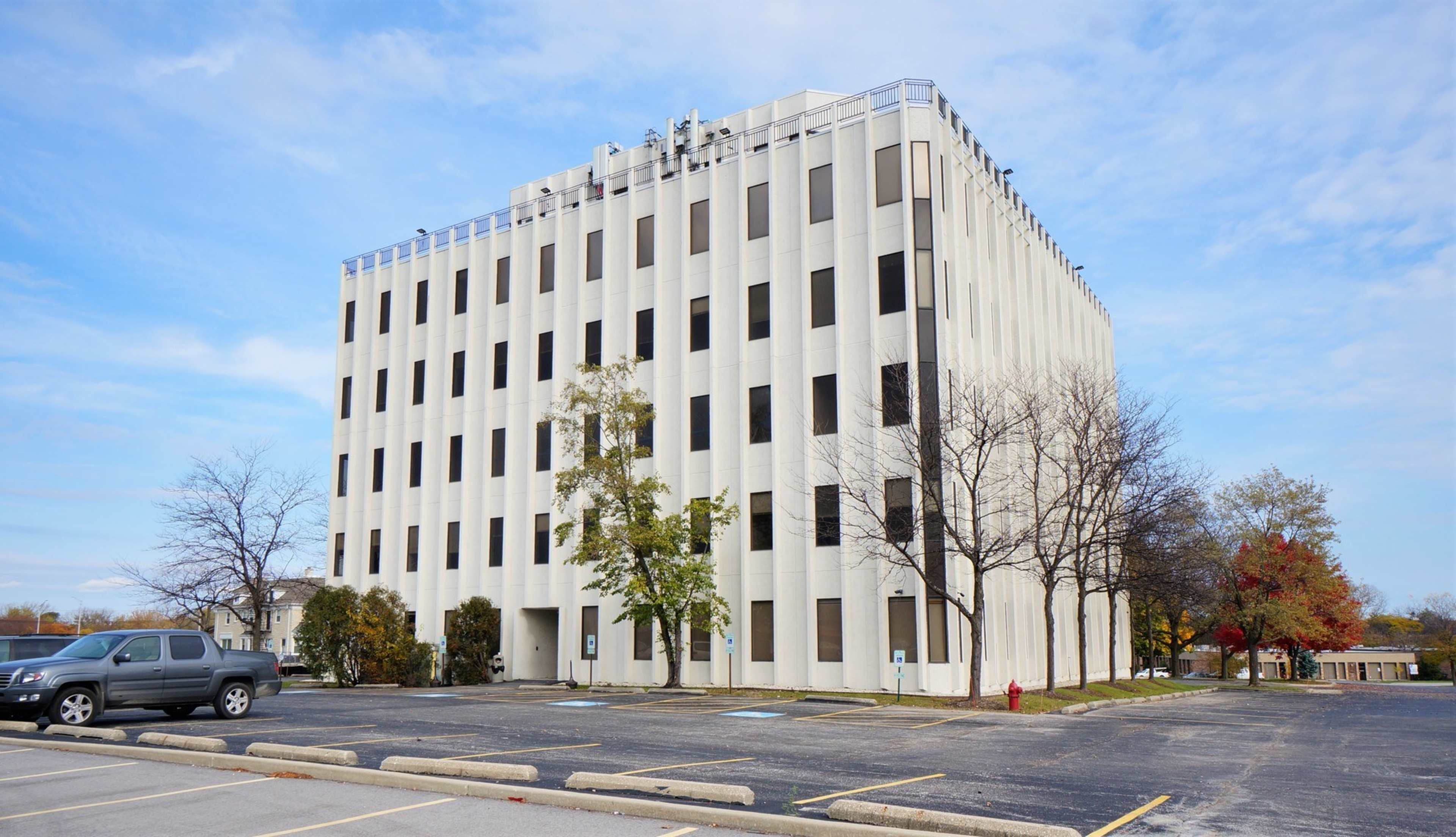 A six-story white office building stands beside a parking lot with sparse trees and a cloudy sky in the background.