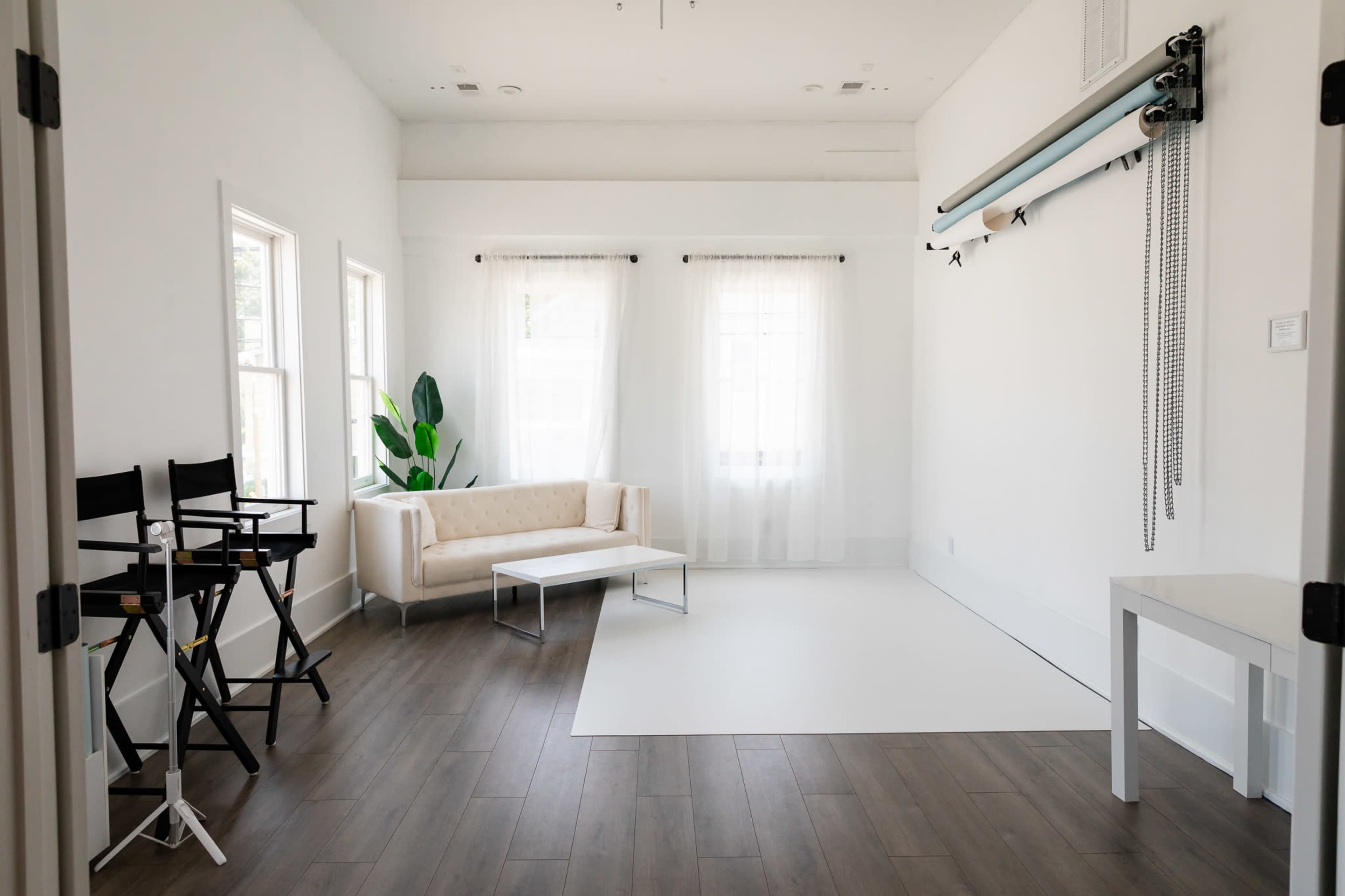 The image shows a bright, minimalistic studio space featuring a beige sofa, a white table, and a backdrop setup against a clean white wall.
