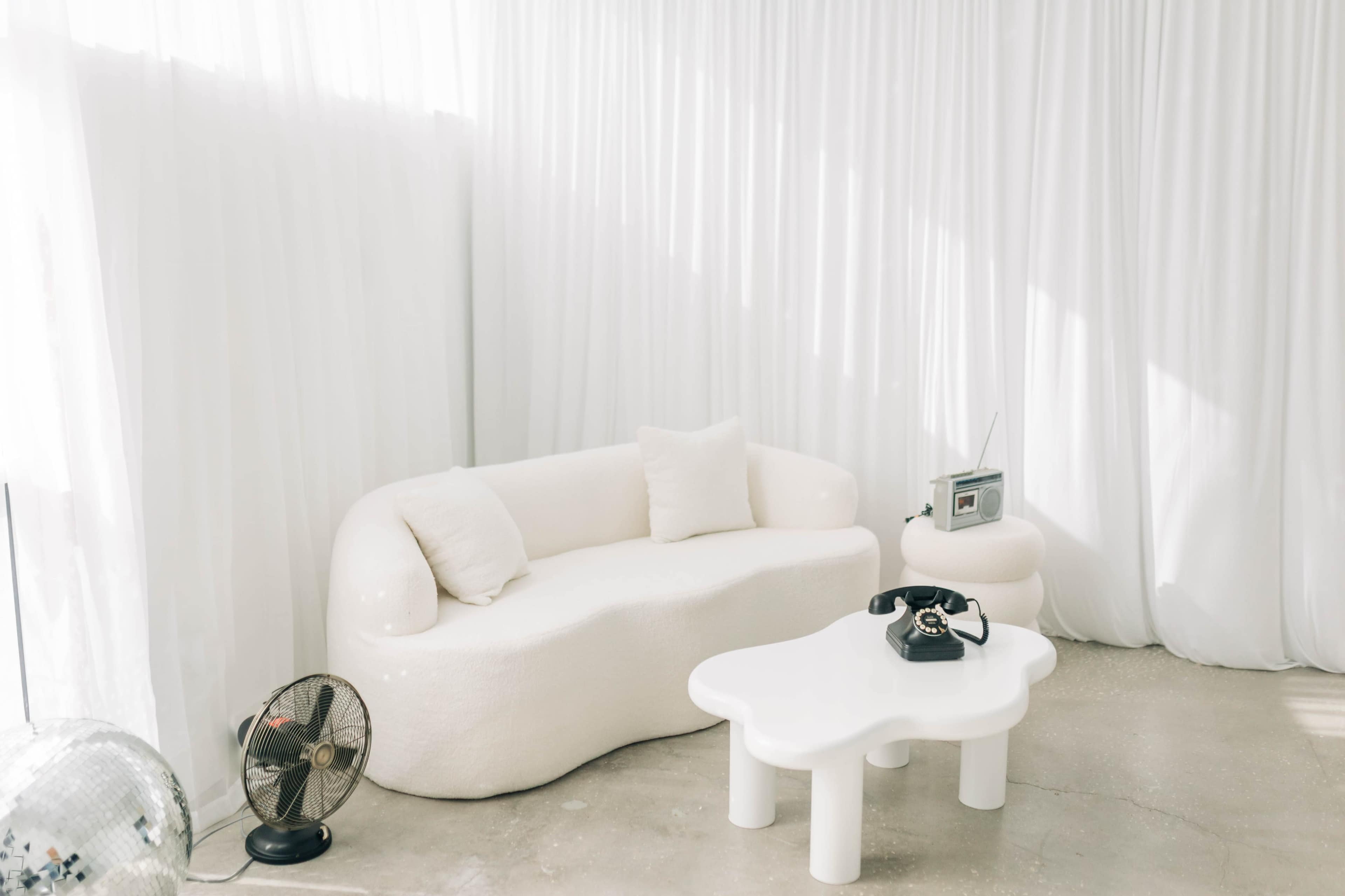 A minimalist white living area features a sculptural sofa, a glossy white coffee table, and a retro-style phone, all against a backdrop of flowing sheer curtains and a small fan.