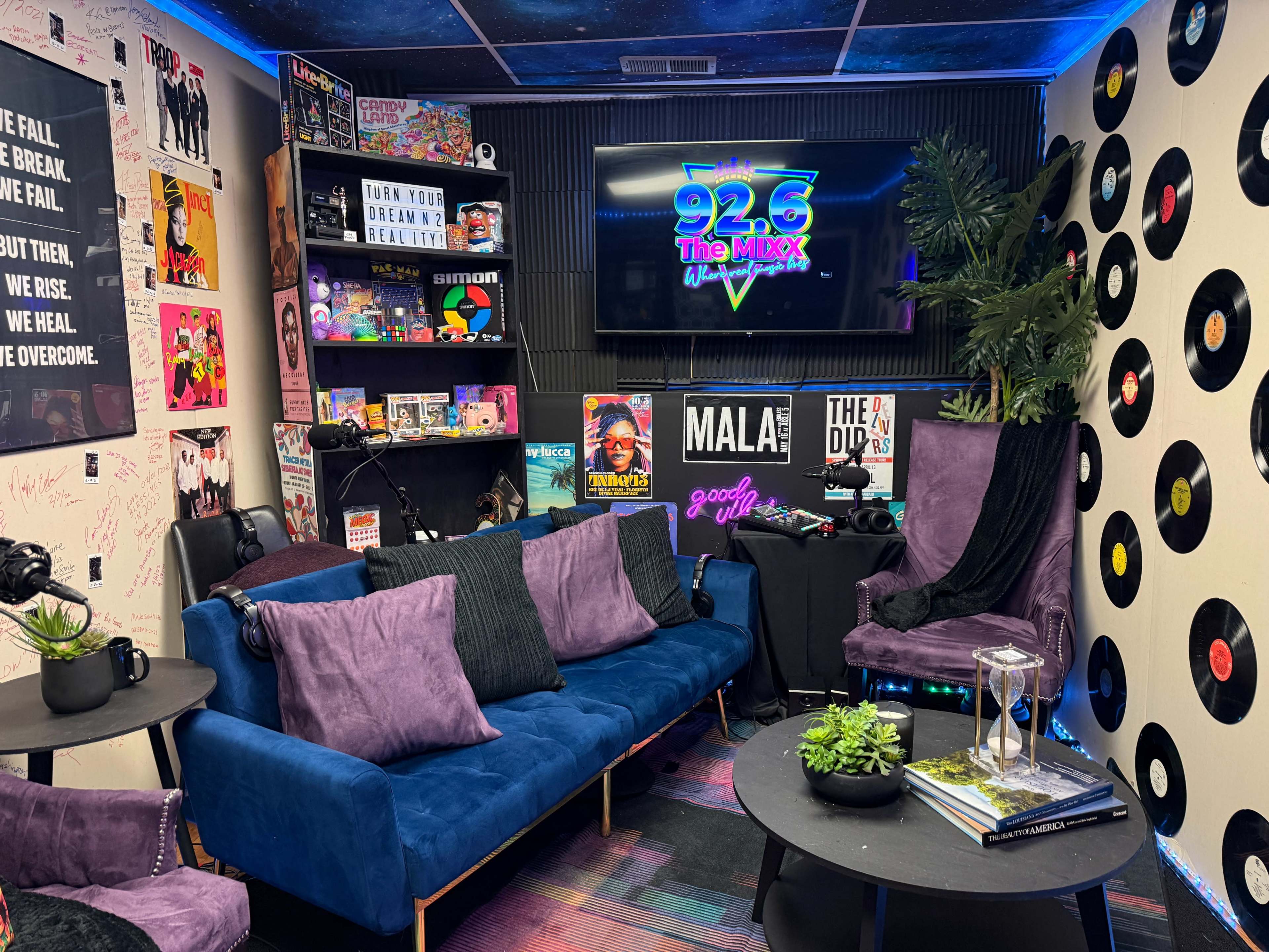 A colorful radio studio features a blue couch, various entertainment memorabilia on the walls, and a large screen displaying the station's logo.
