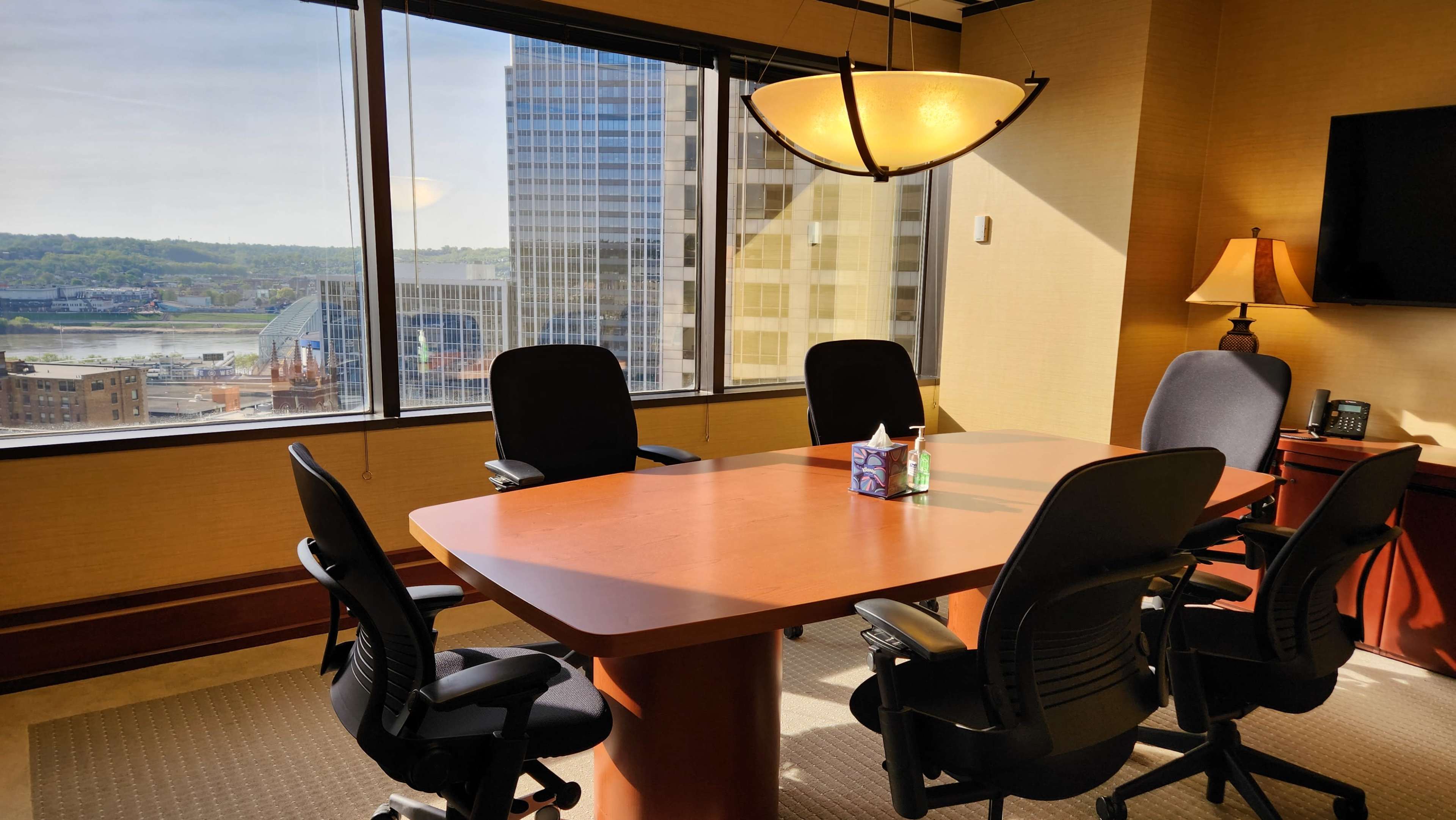 A conference room features a large wooden table surrounded by six black chairs, with large windows showcasing a cityscape and river view.