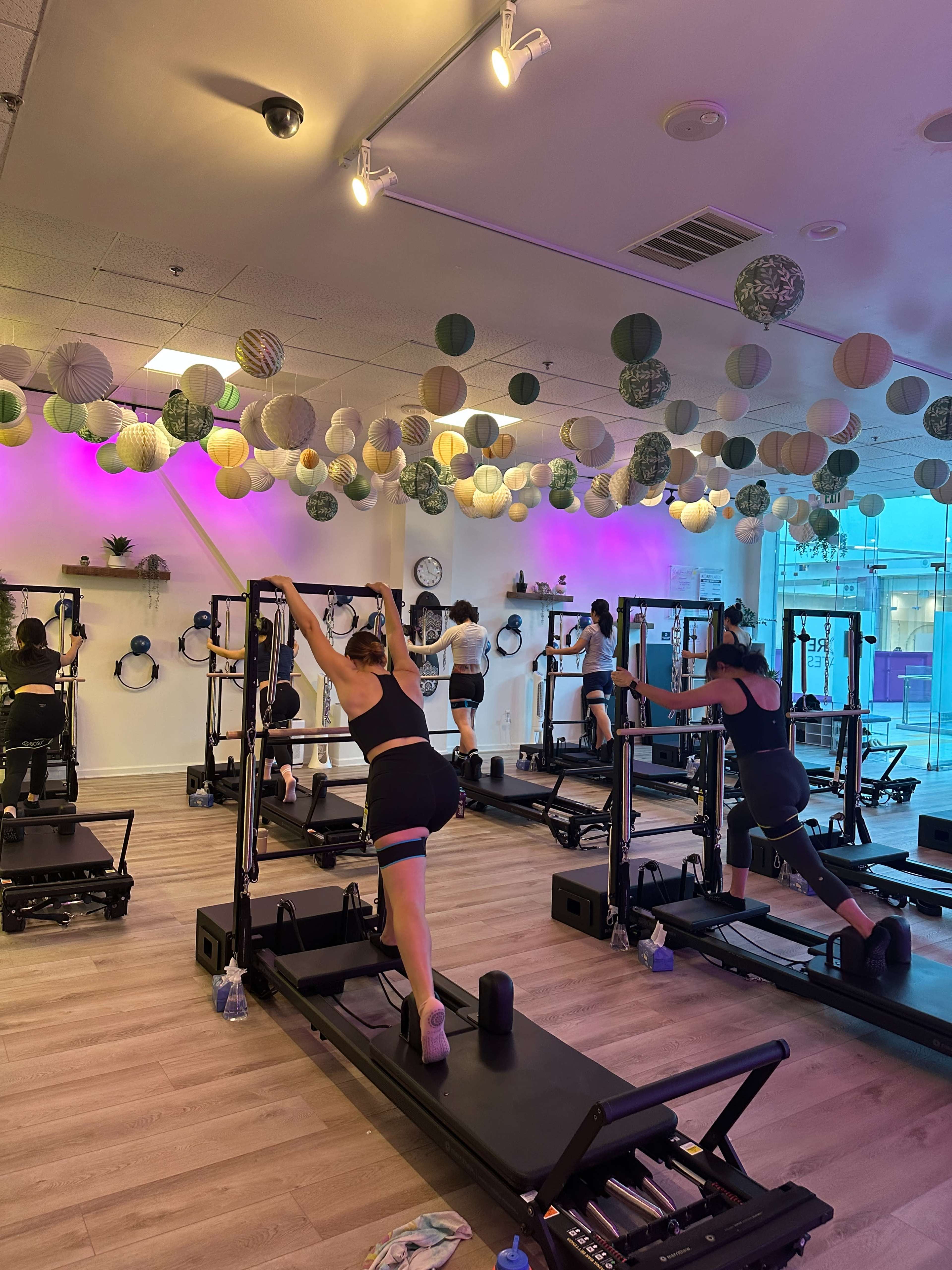 A group of individuals is engaged in a fitness class using reformer machines in a brightly lit studio adorned with colorful hanging decorations.