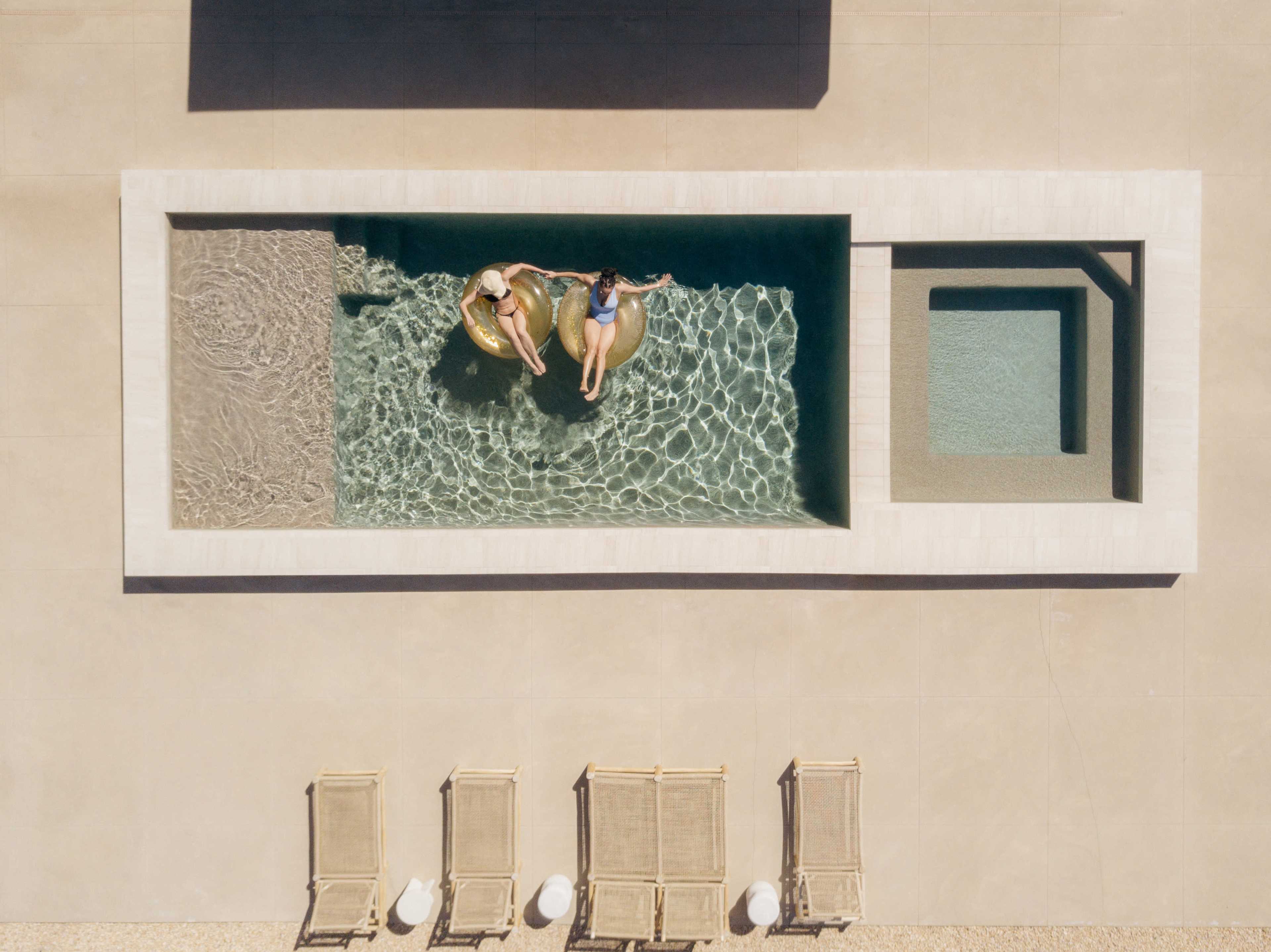 Two individuals relax on floatation devices in a rectangular swimming pool, surrounded by lounge chairs and a spa area.