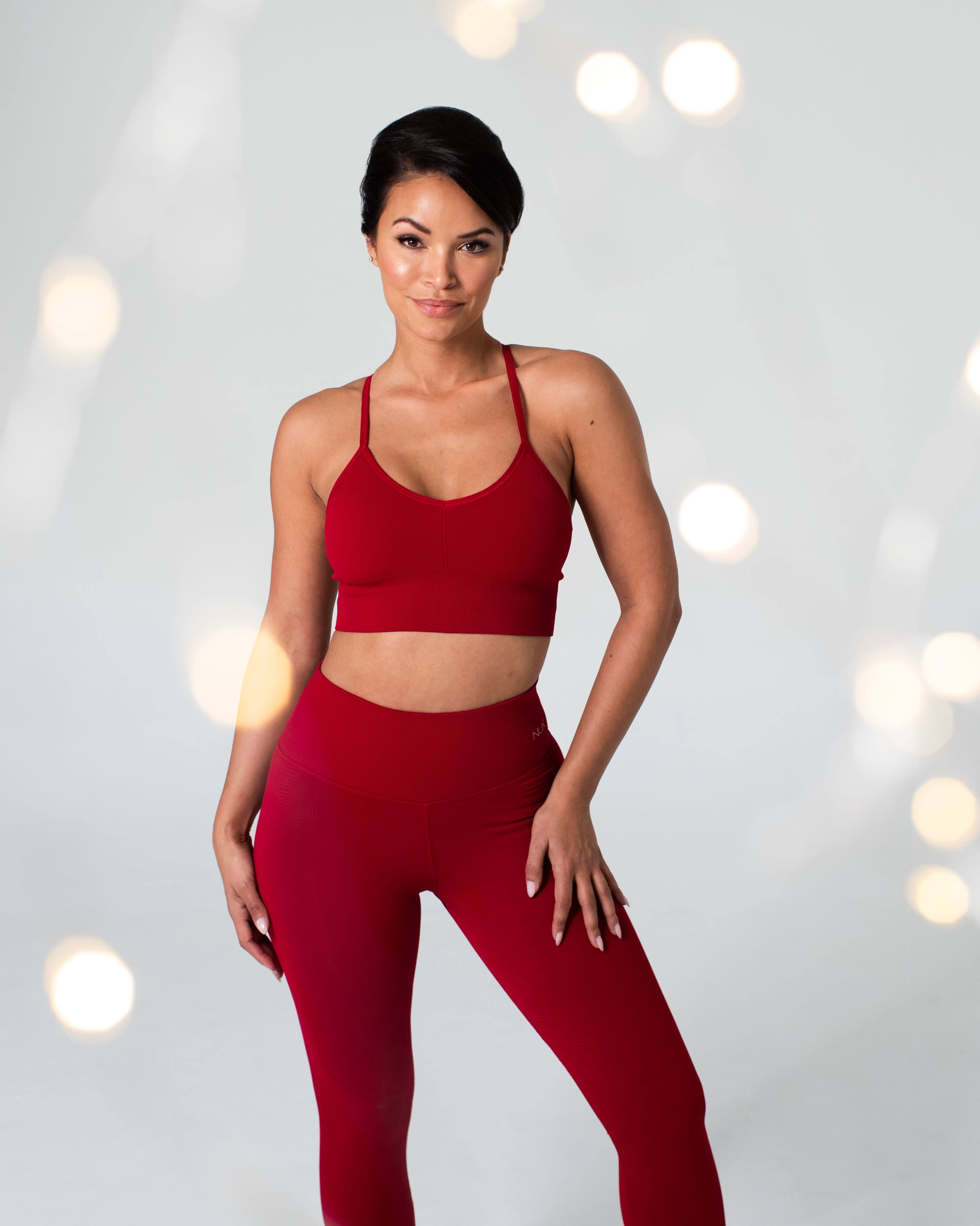A woman stands confidently in a red sports bra and matching leggings against a light gray background.