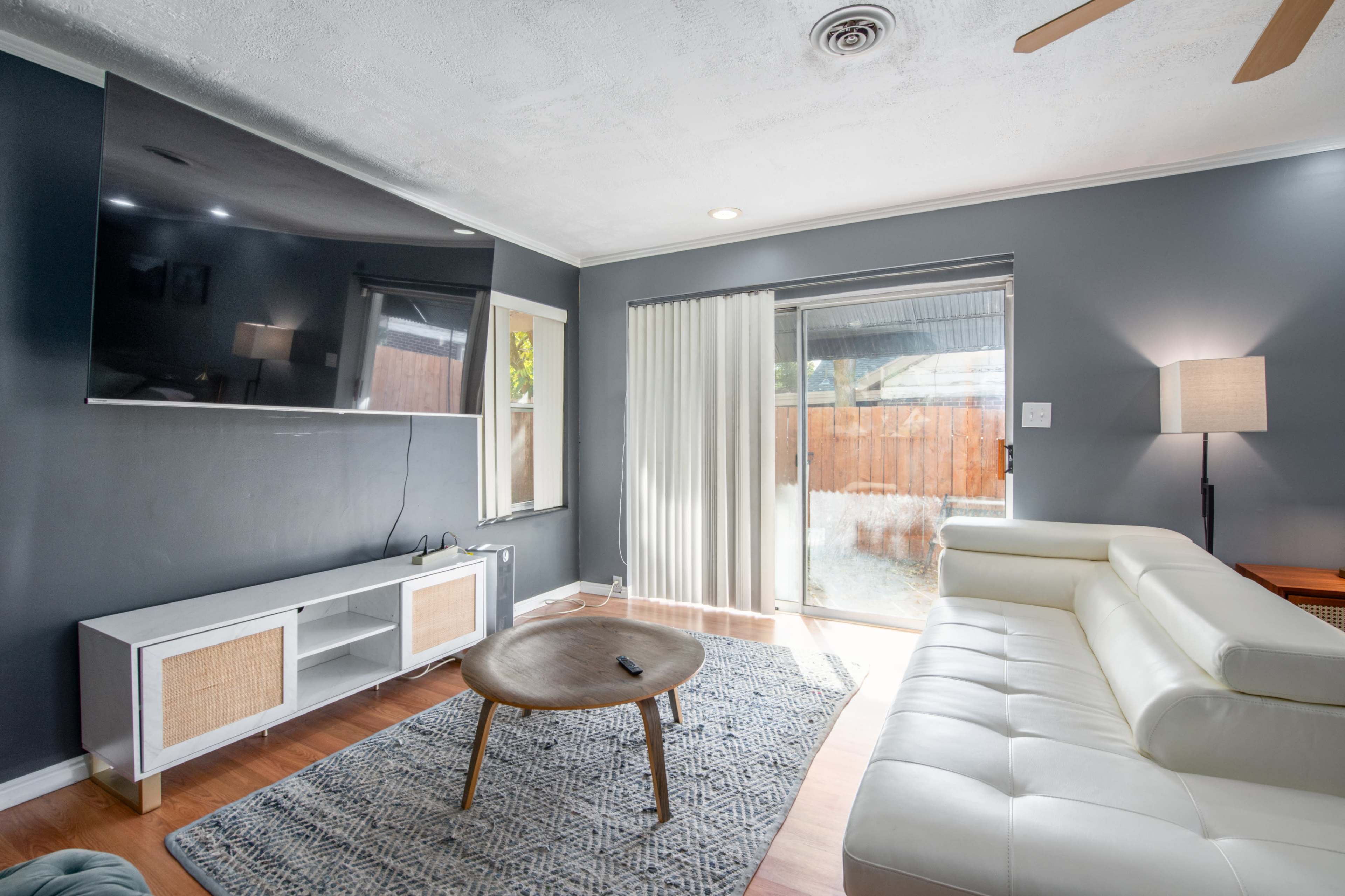 A living room with a white sectional sofa, a wooden coffee table, a wall-mounted television, and sliding glass doors leading to an outdoor patio.