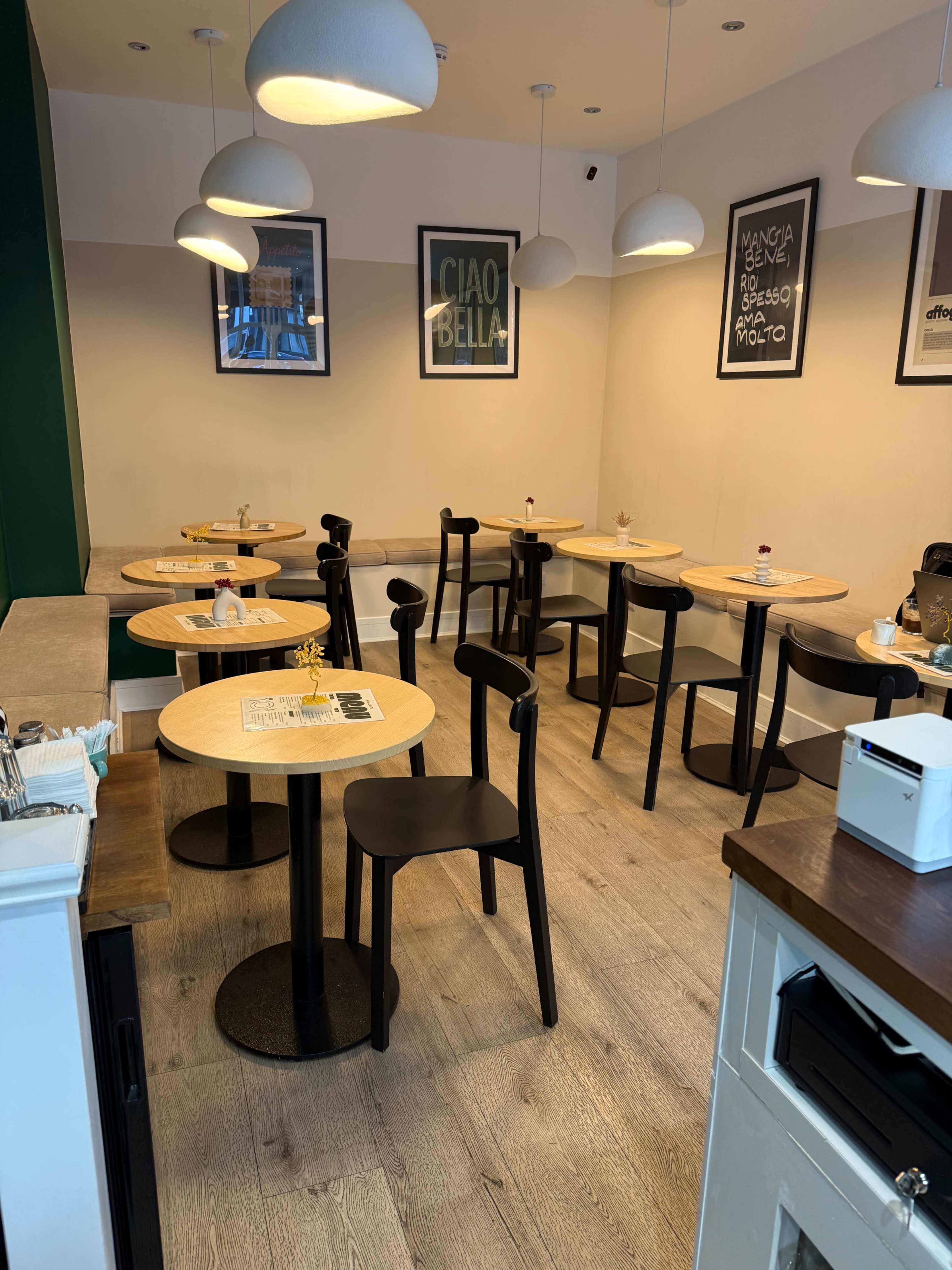 The image shows a modern café interior with wooden tables and black chairs arranged neatly, and light fixtures hanging from the ceiling.