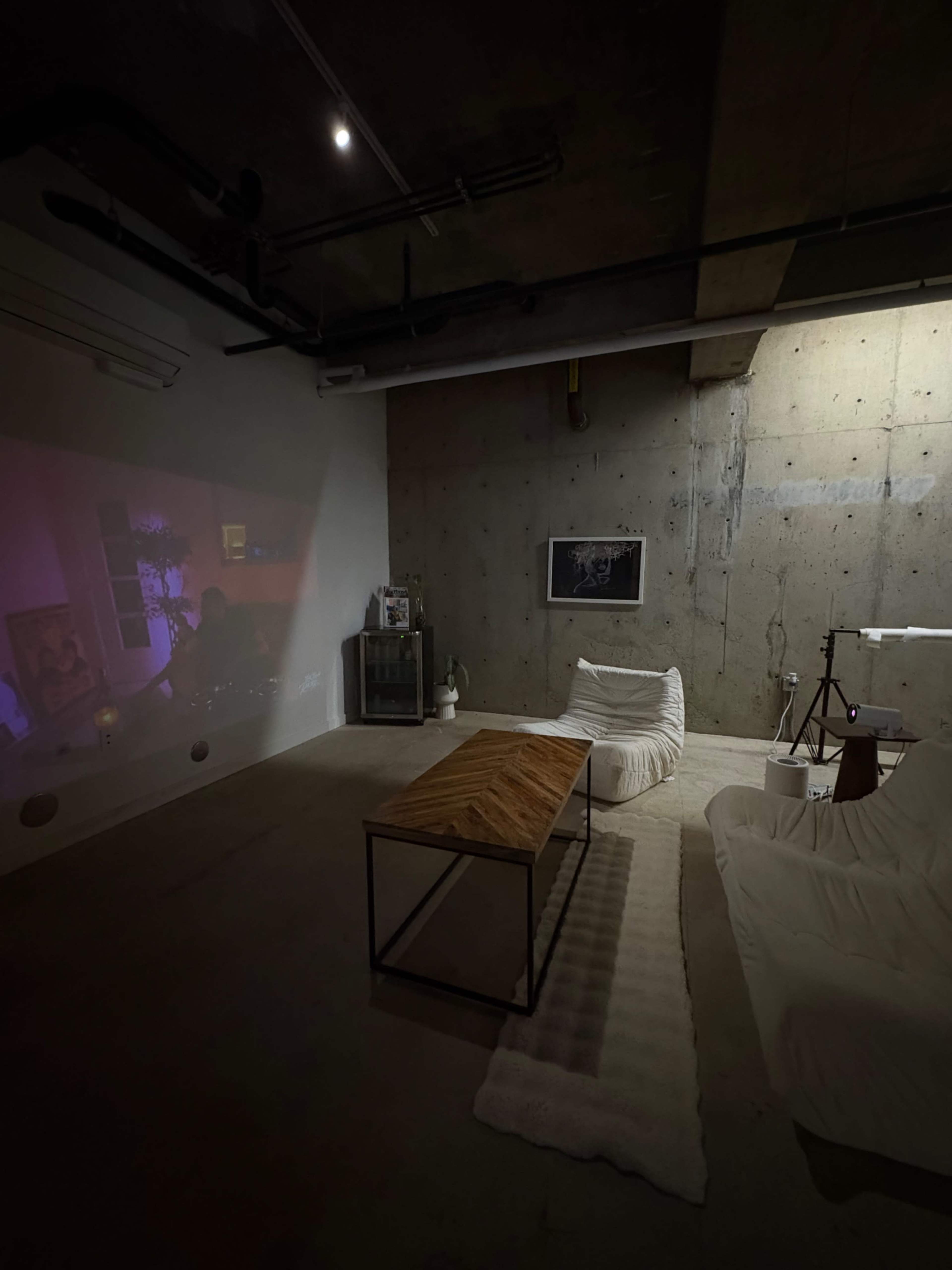 The image shows a dimly lit industrial-style room with a concrete wall, a wooden coffee table, two white beanbag chairs, and a projection on the wall displaying a scene with soft lighting.