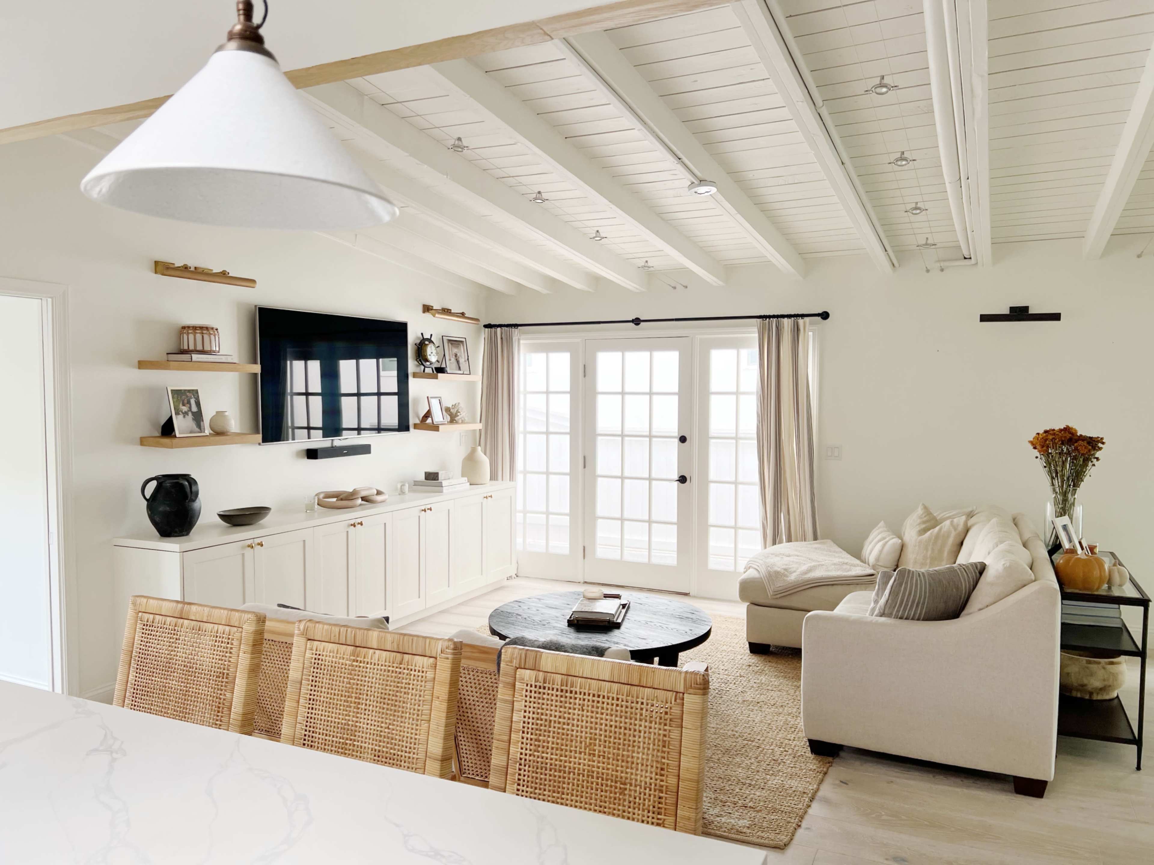 A bright living room features a cream sectional sofa, a round coffee table, and a large TV mounted on a white wall with built-in shelves.