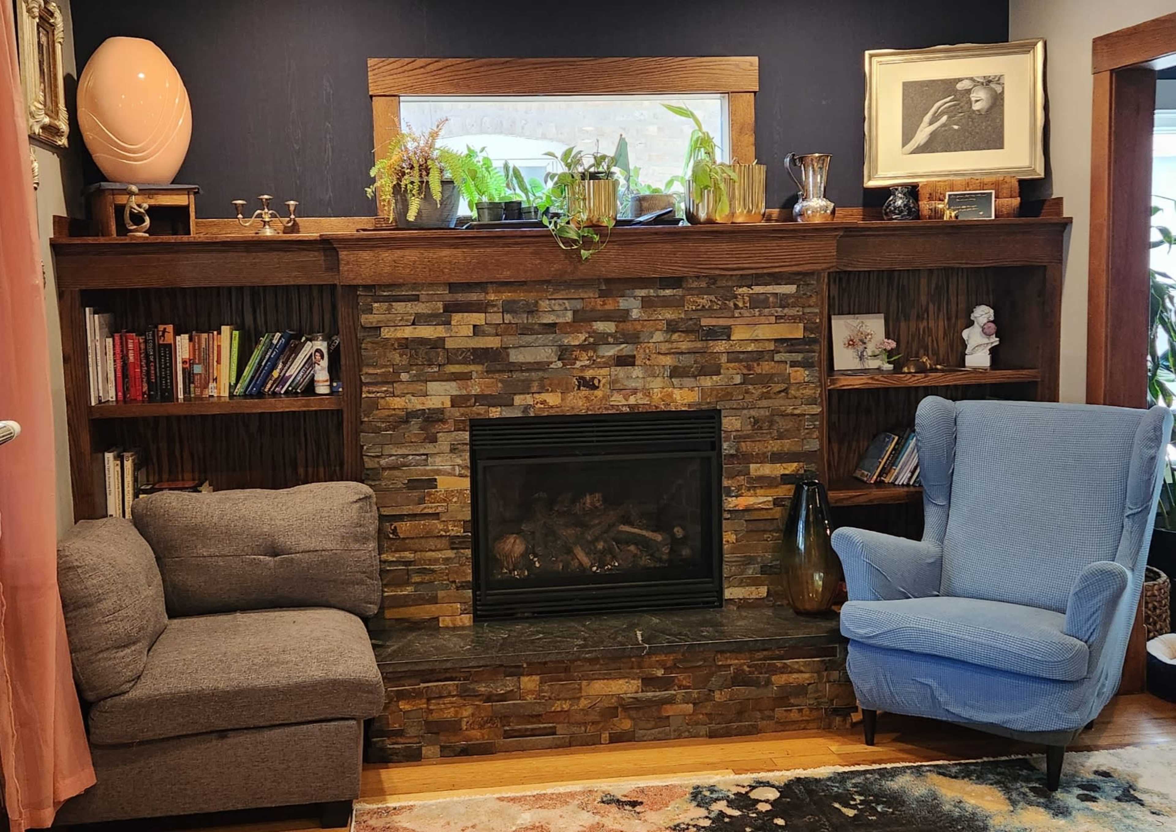 The image depicts a cozy living room with a stone fireplace surrounded by wooden shelves filled with books and decorative items, alongside a blue armchair and a gray sofa.