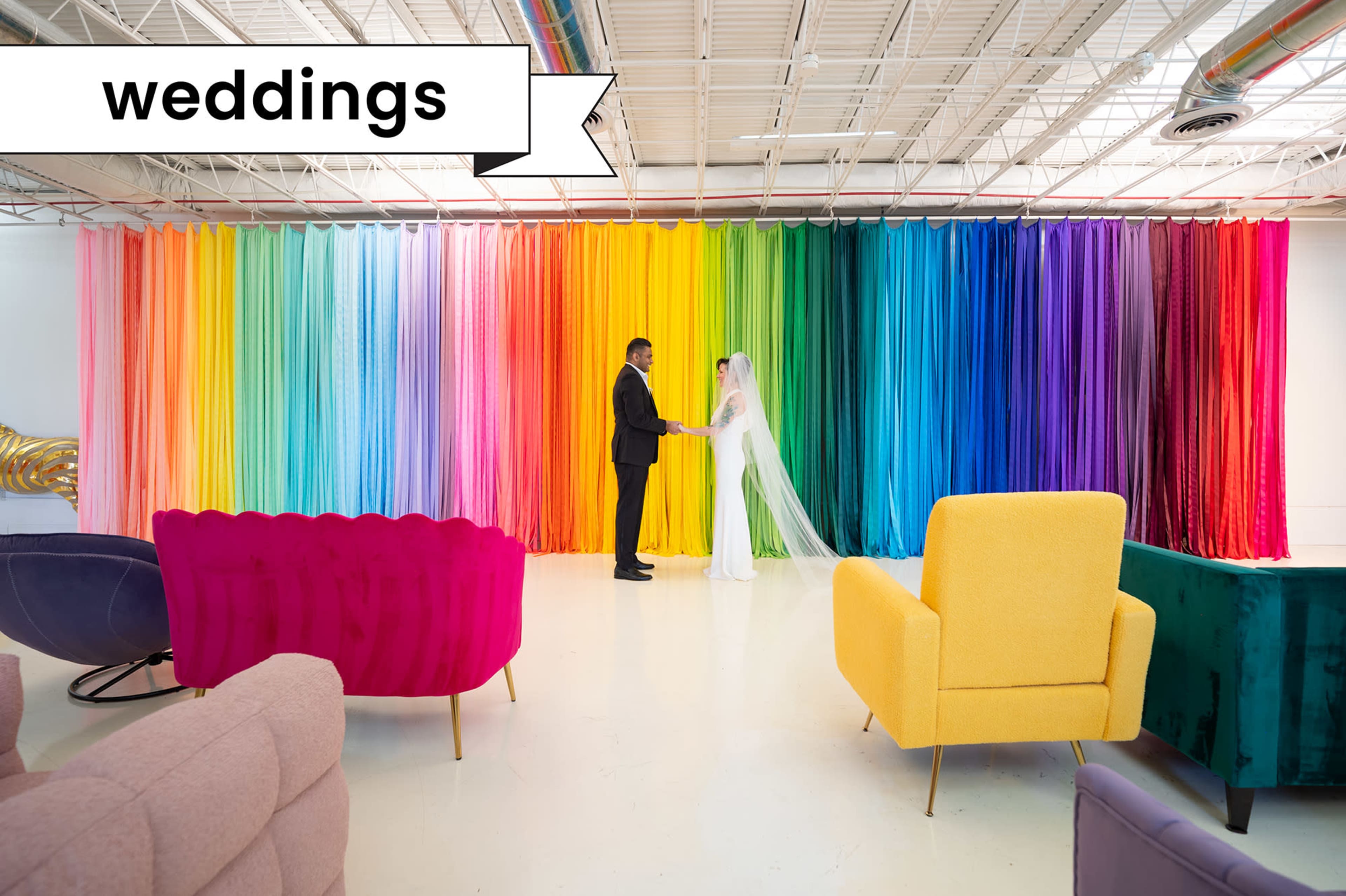 A couple stands facing each other in front of a vibrant, multicolored curtain backdrop, with stylish seating in the foreground.