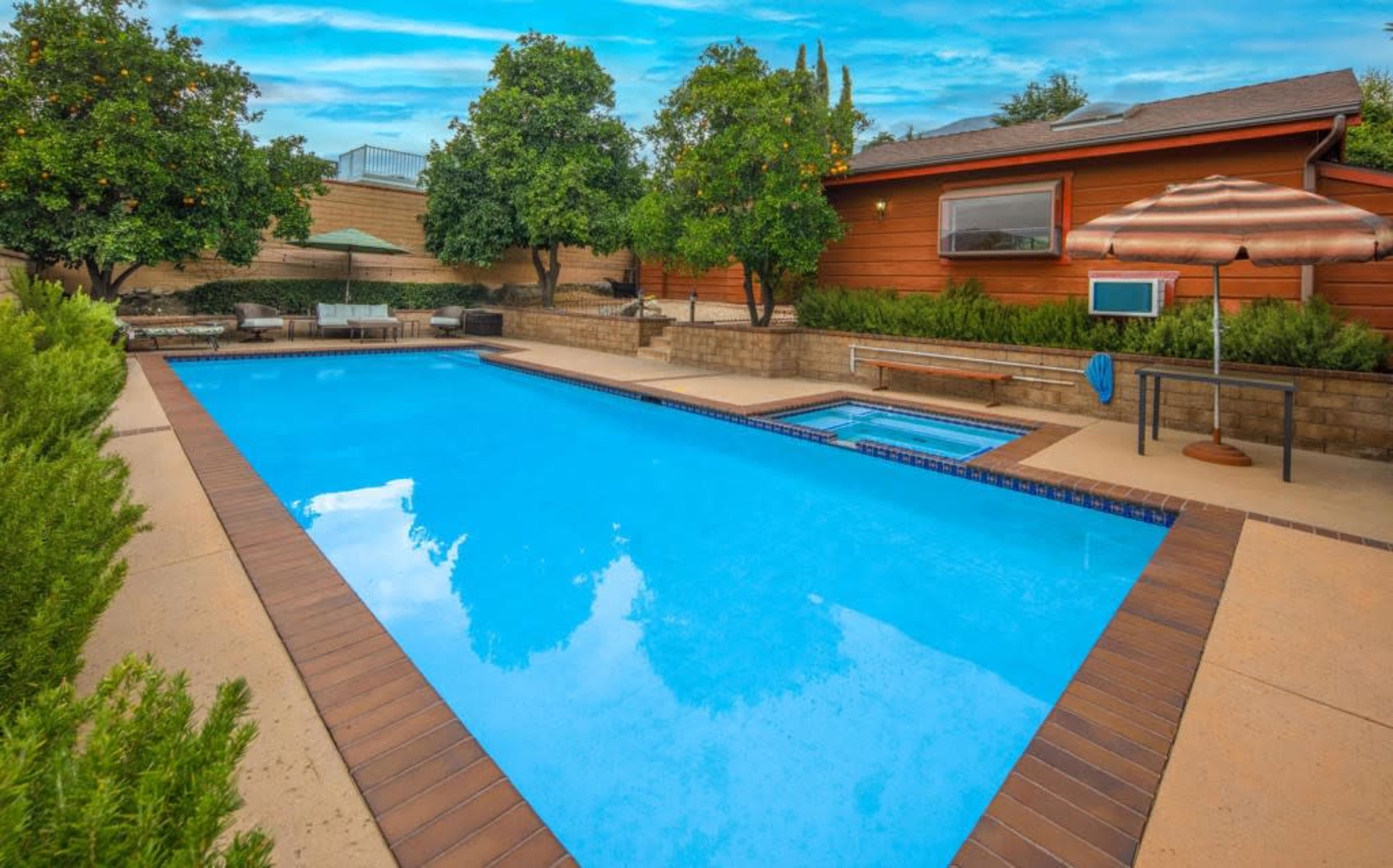 A residential backyard featuring a blue swimming pool with a hot tub, surrounded by green trees and a house in the background.
